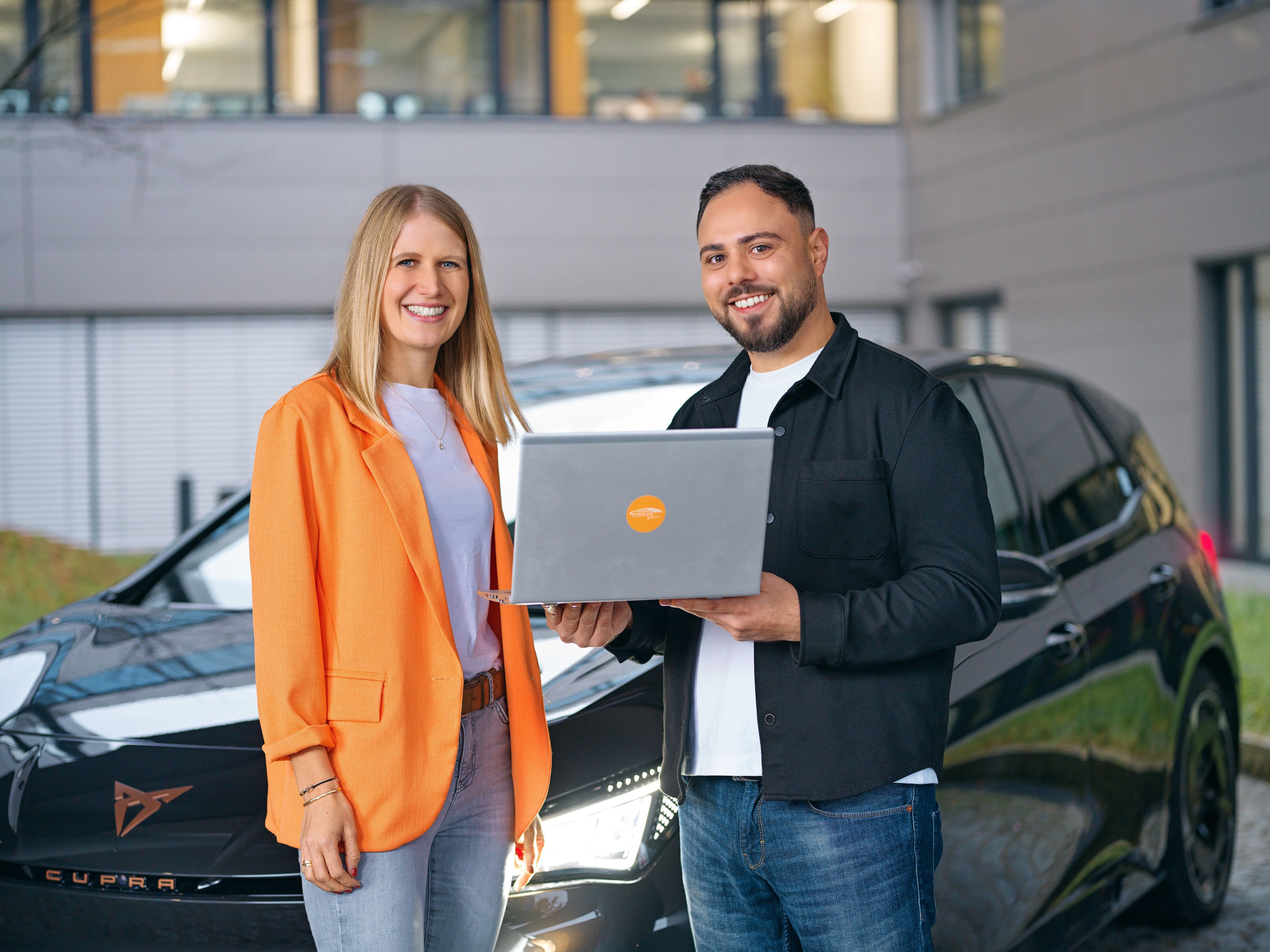 Two people are smiling in front of a CUPRA, a laptop in their hands.