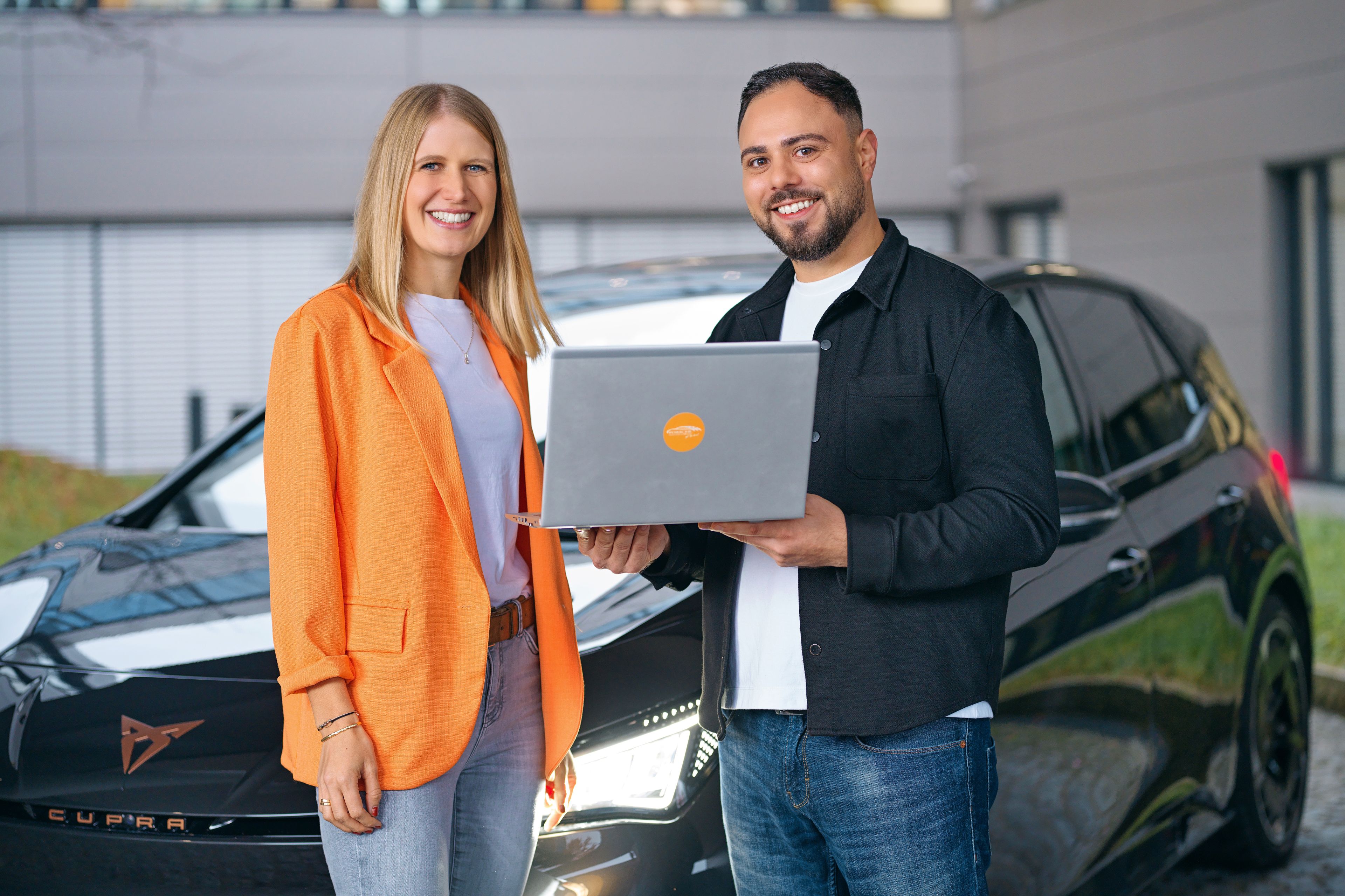 Two people are smiling in front of a CUPRA, a laptop in their hands.