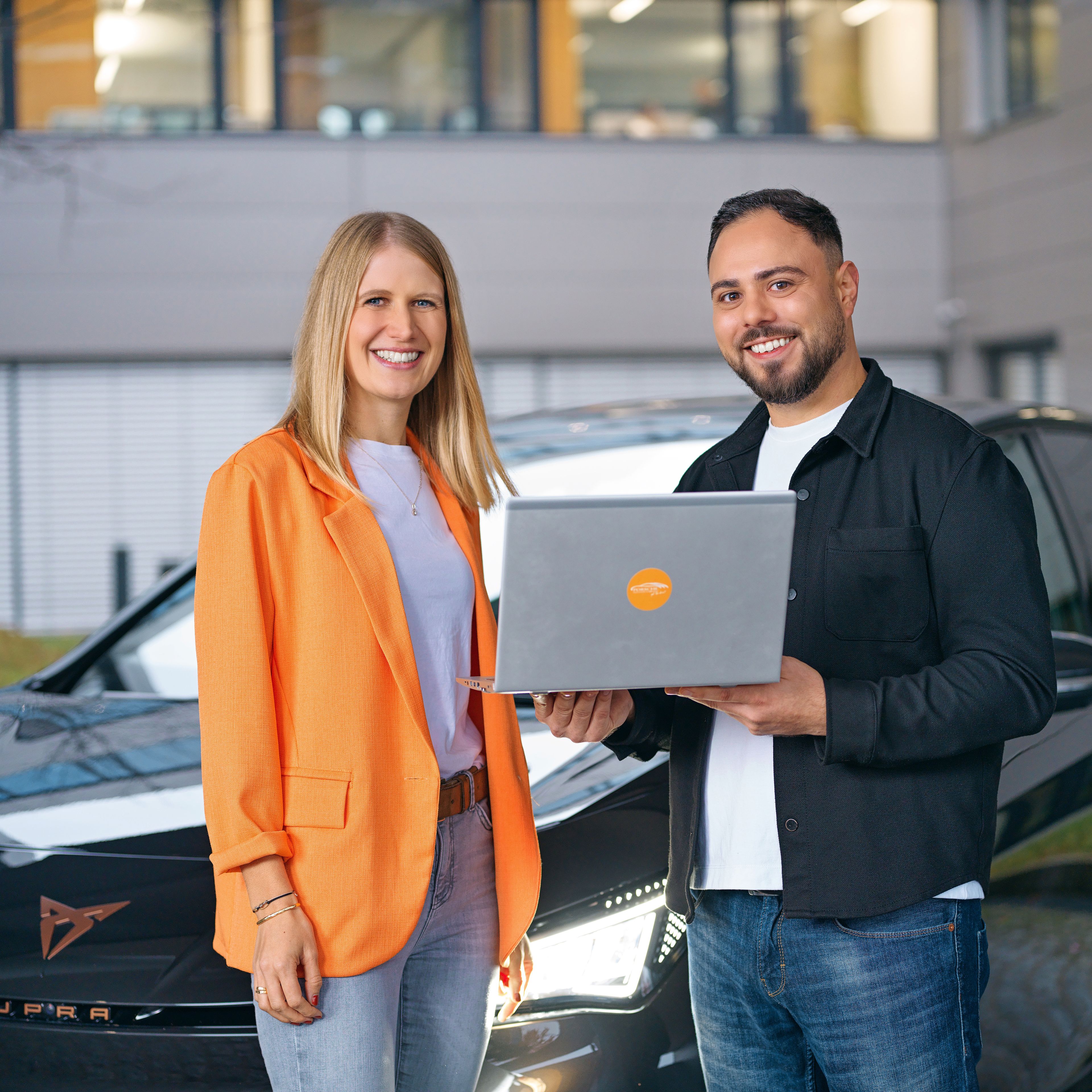 Two people are smiling in front of a CUPRA, a laptop in their hands.