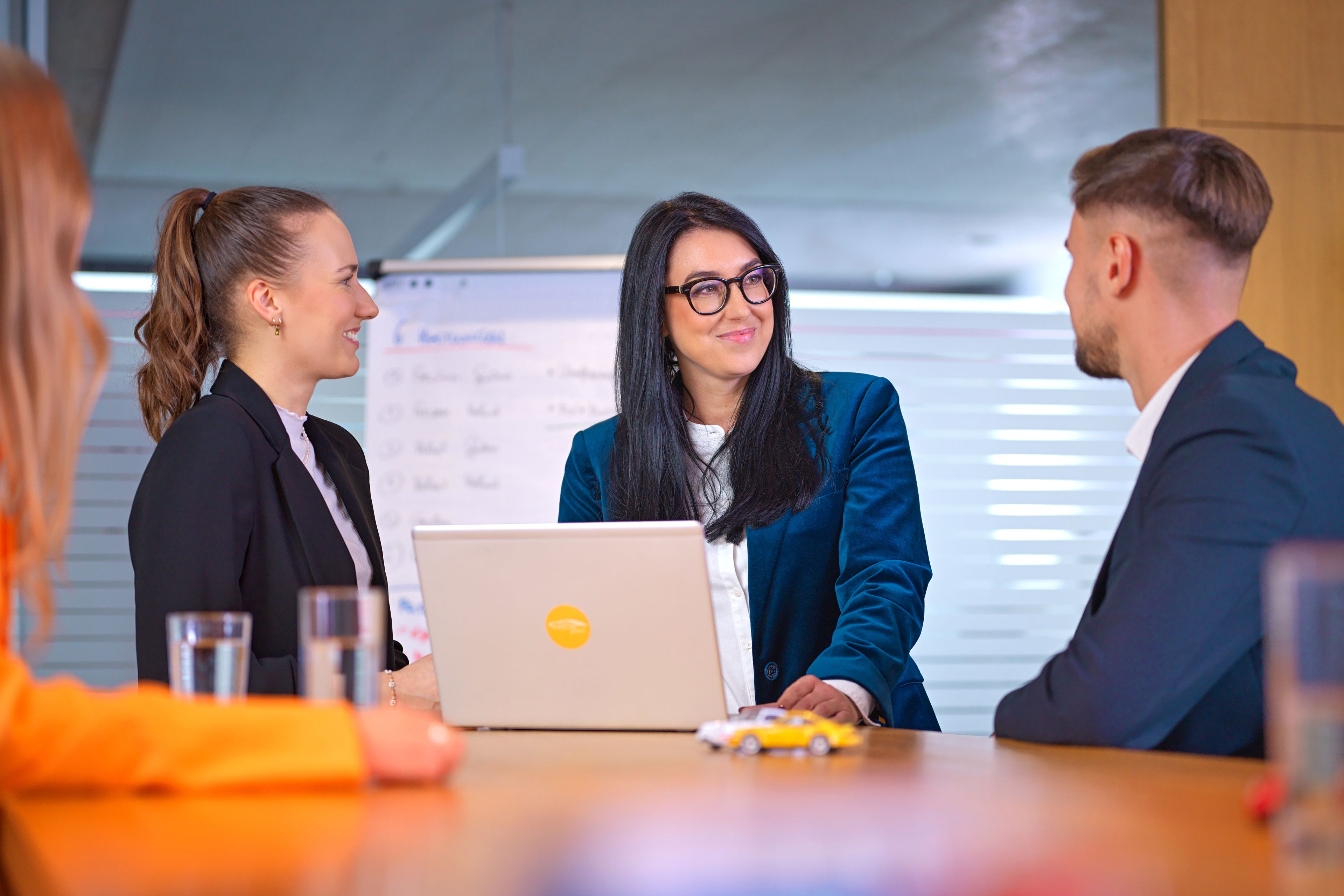 Hárman, két hölgy és egy úriember beszélgetnek egy kávézóban, mögöttük fekete-fehér fotók régi autóversenyekről.