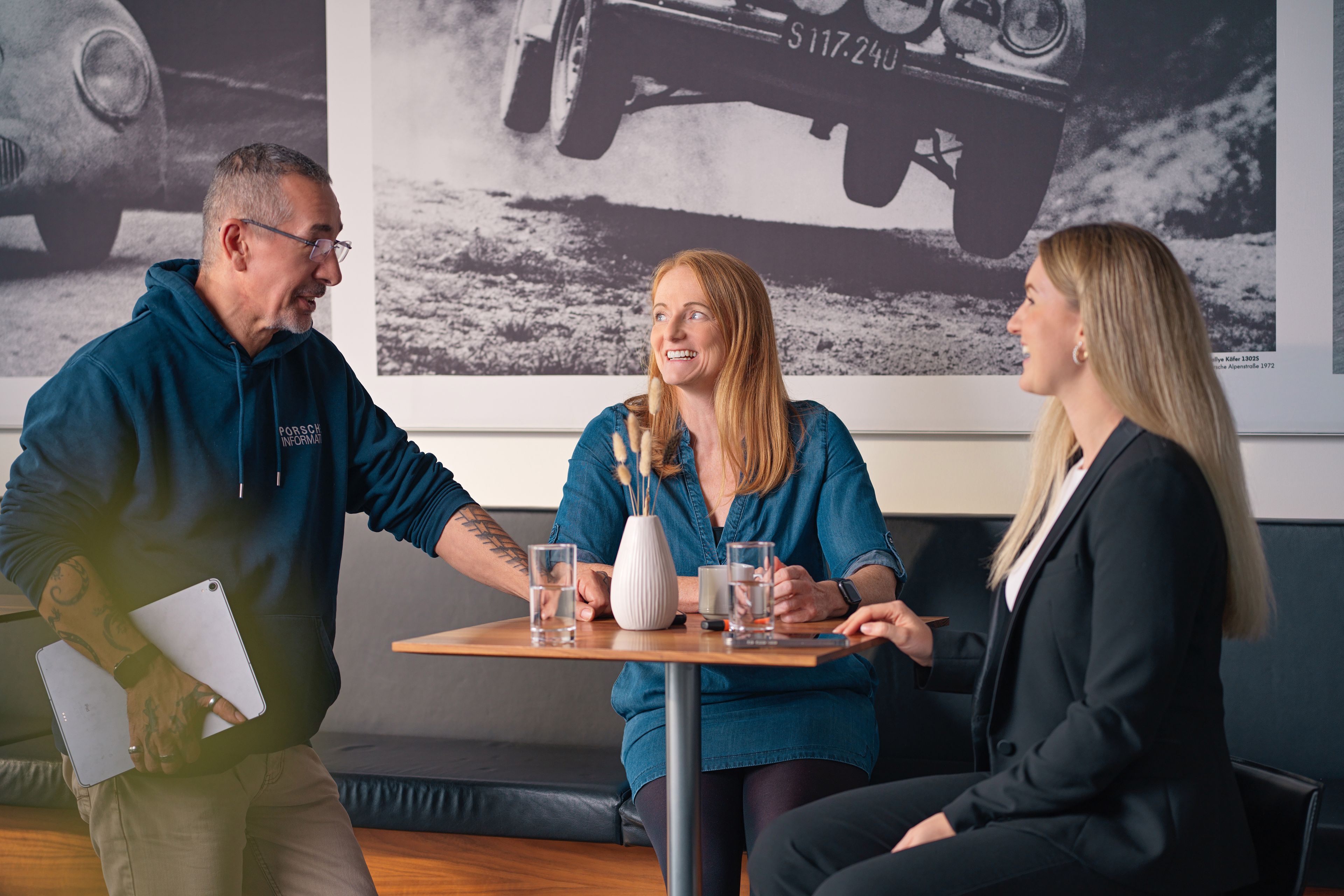 Three people, two ladies and a gentleman, are talking in a café, with black and white photos of old car races behind them.