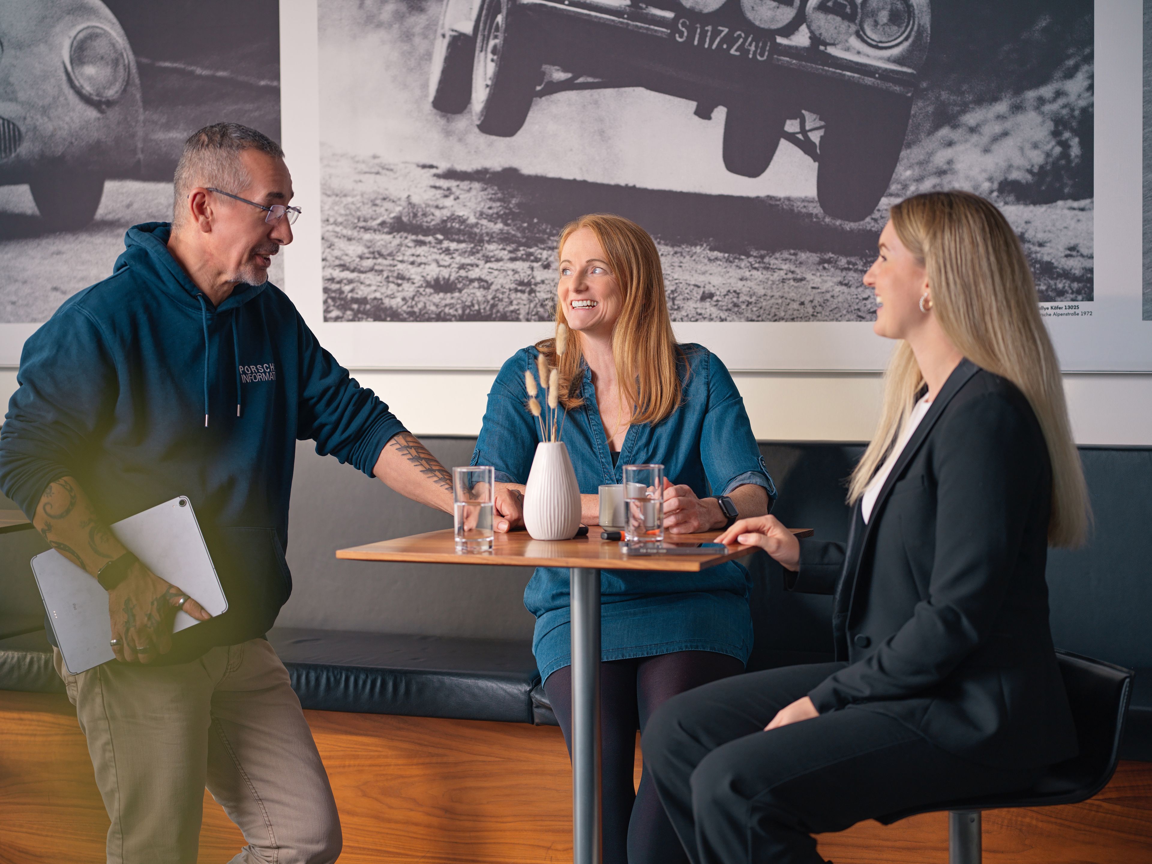 Three people, two ladies and a gentleman, are talking in a café, with black and white photos of old car races behind them.