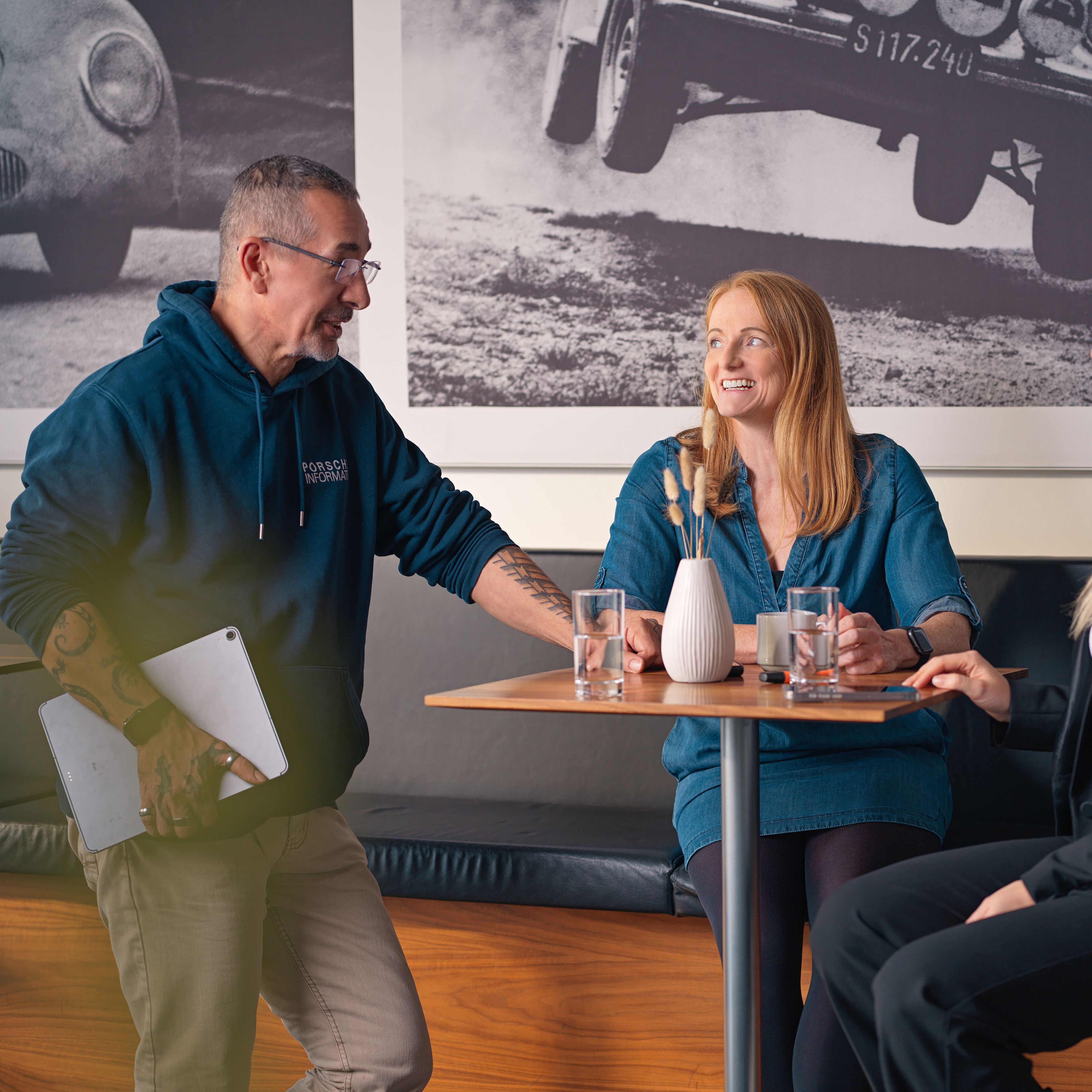 Three people, two ladies and a gentleman, are talking in a café, with black and white photos of old car races behind them.