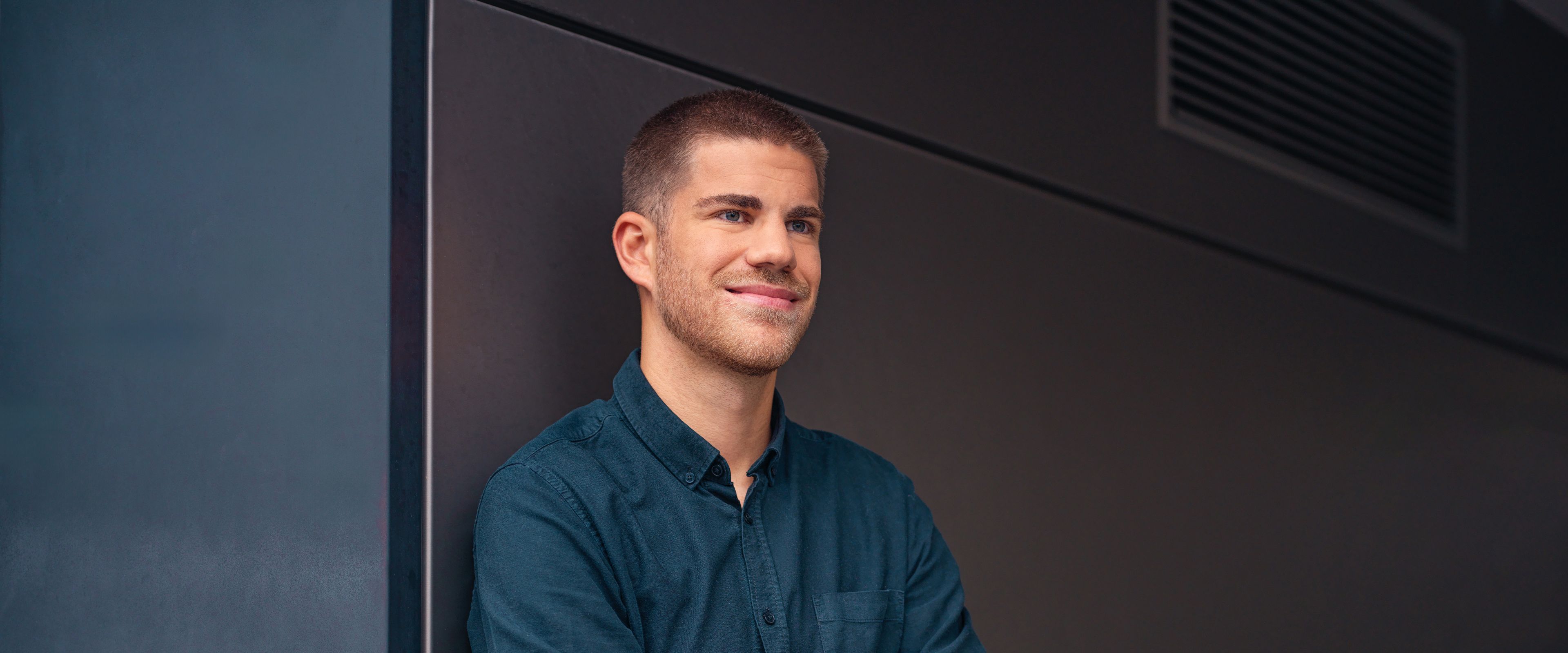 A young man leans against a black wall with his hands in a carbaton.