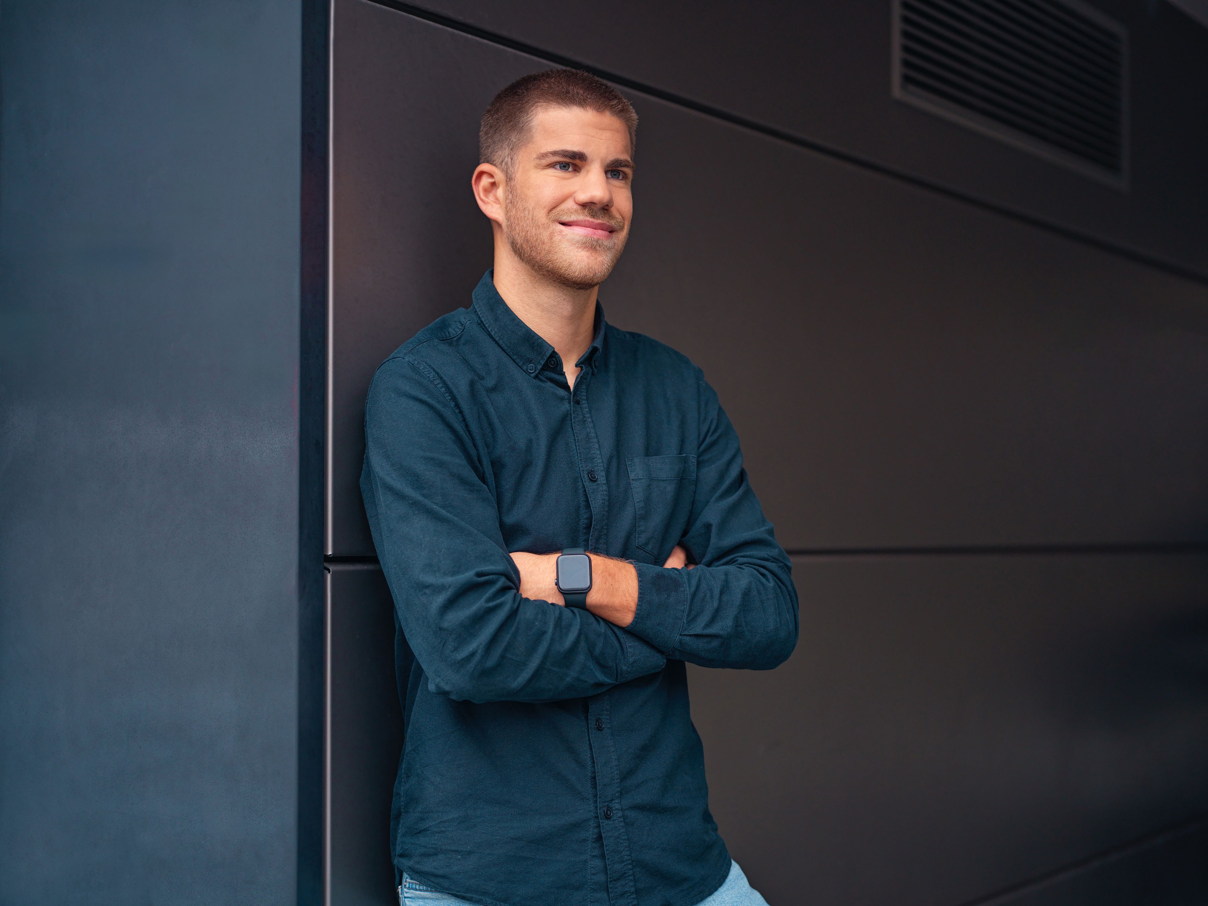 A young man leans against a black wall with his hands in a carbaton.