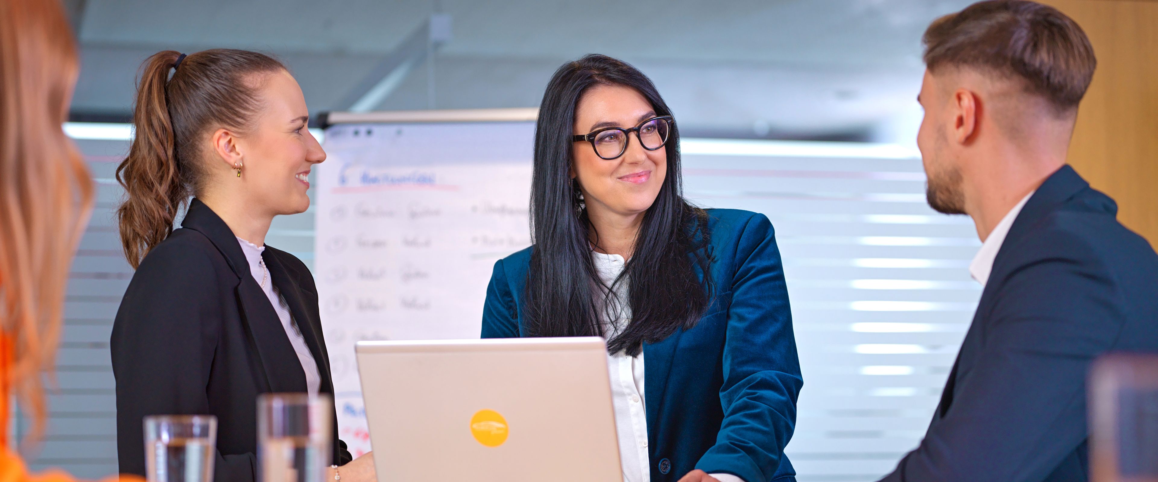 Three people, two ladies and a gentleman, are talking in a café, with black and white photos of old car races behind them.