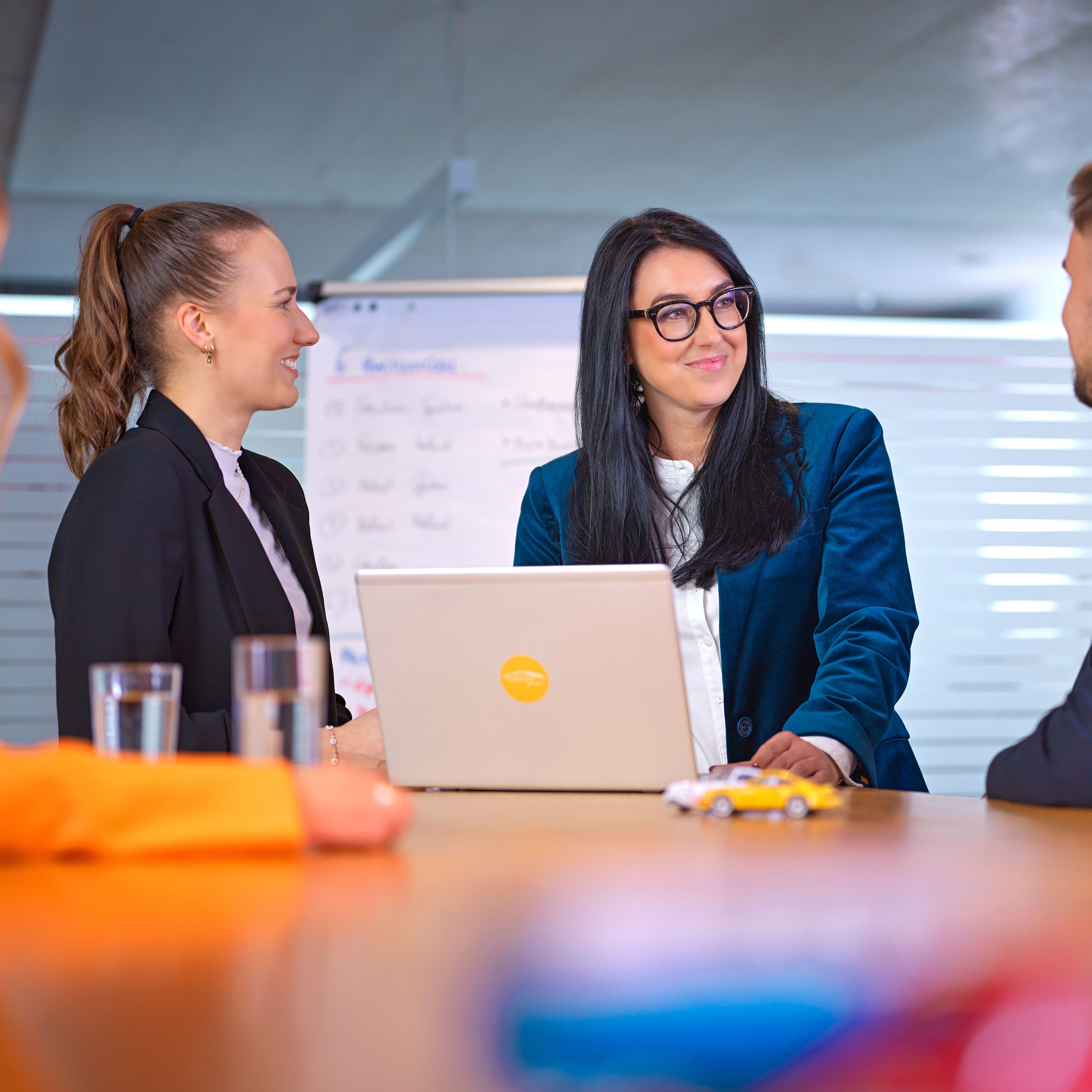 Three people, two ladies and a gentleman, are talking in a café, with black and white photos of old car races behind them.