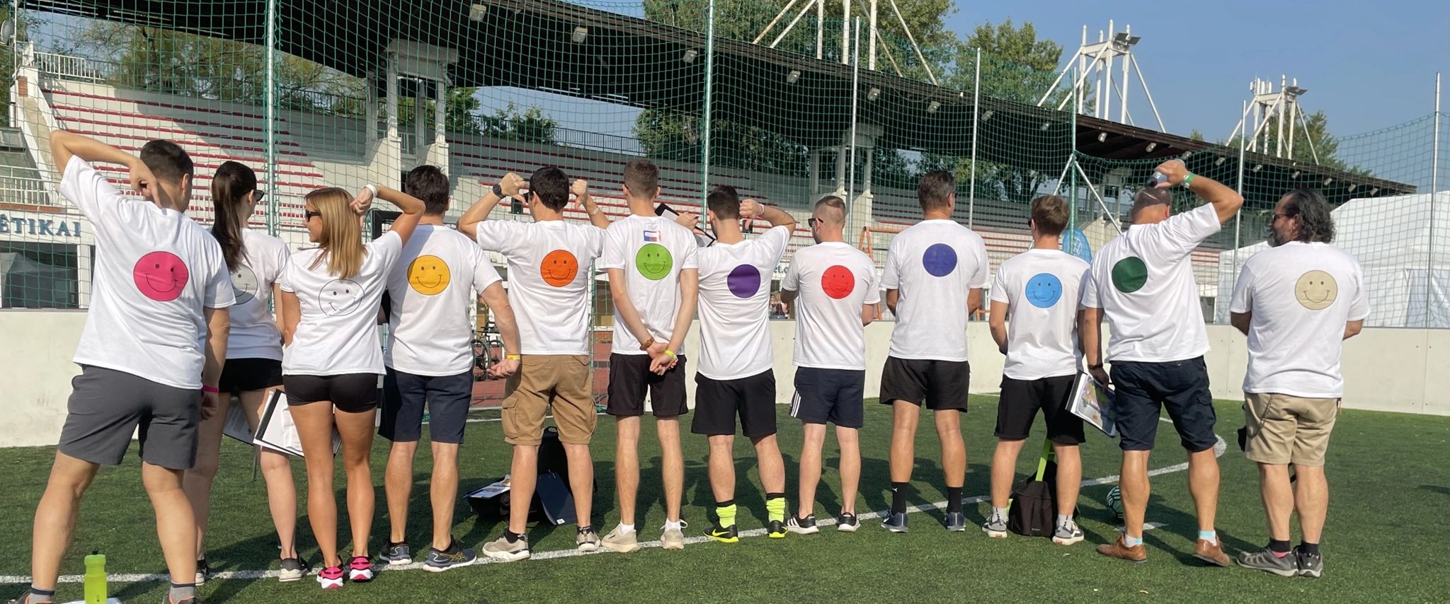A group stands with their backs on a sports field, their T-shirts have colorful smileys.