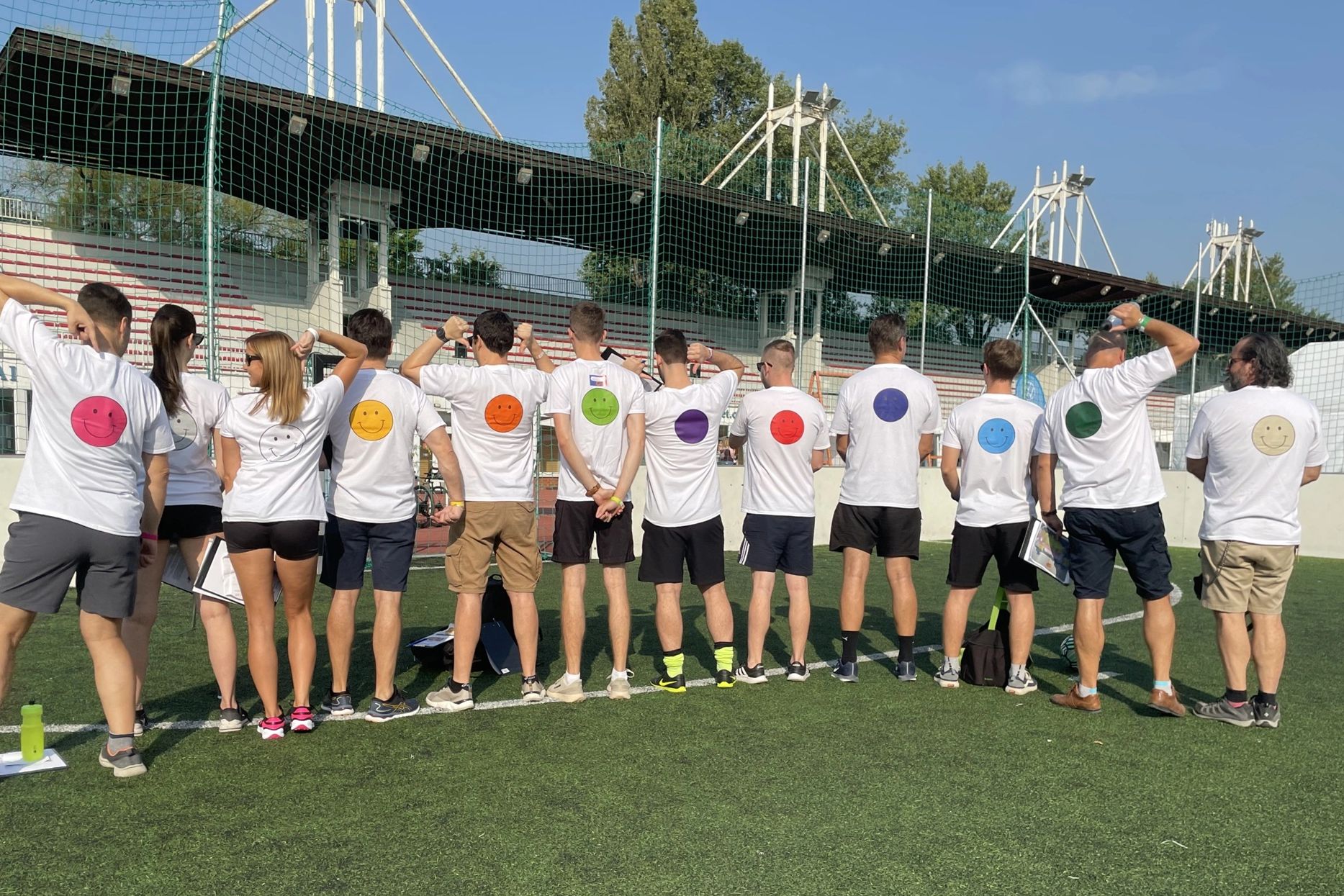 A group stands with their backs on a sports field, their T-shirts have colorful smileys.