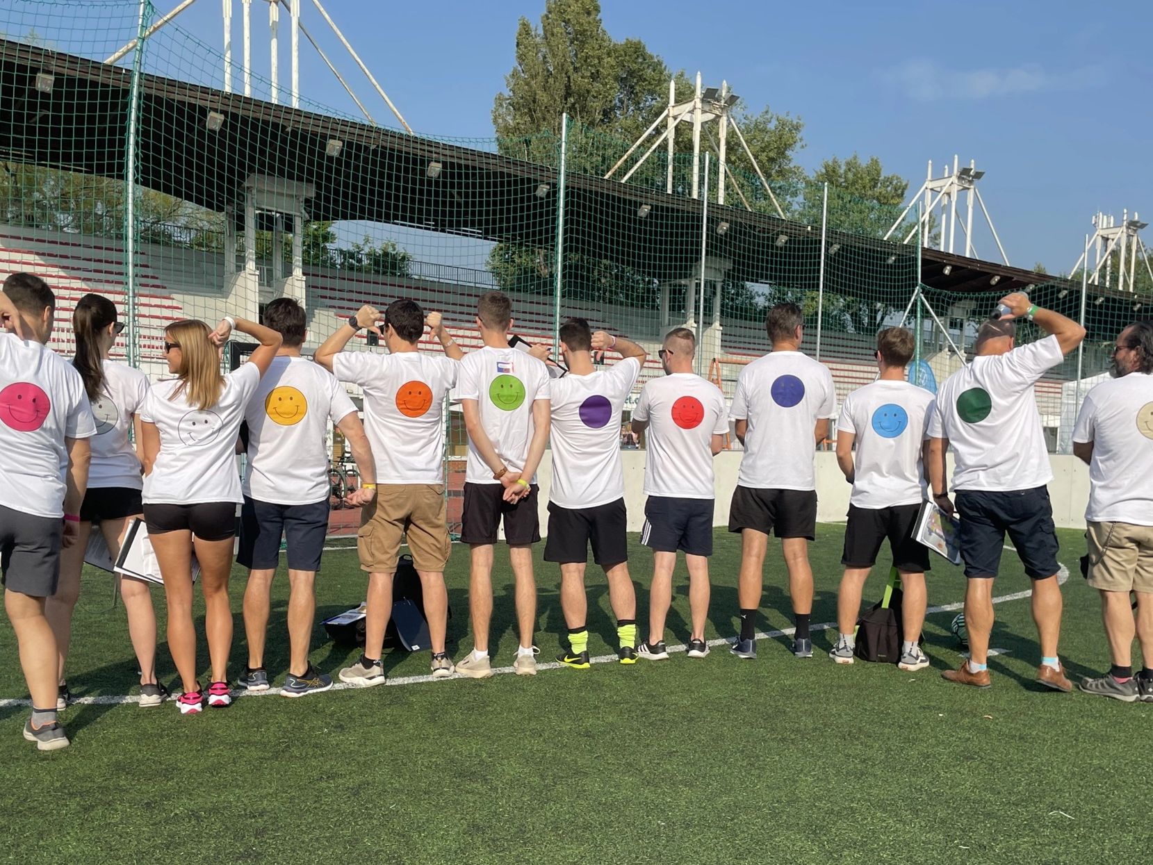 A group stands with their backs on a sports field, their T-shirts have colorful smileys.