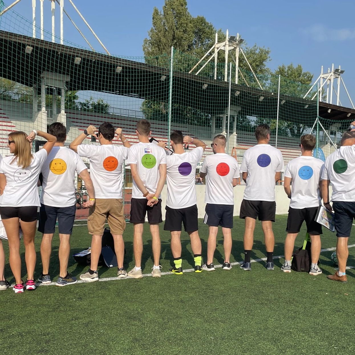 A group stands with their backs on a sports field, their T-shirts have colorful smileys.