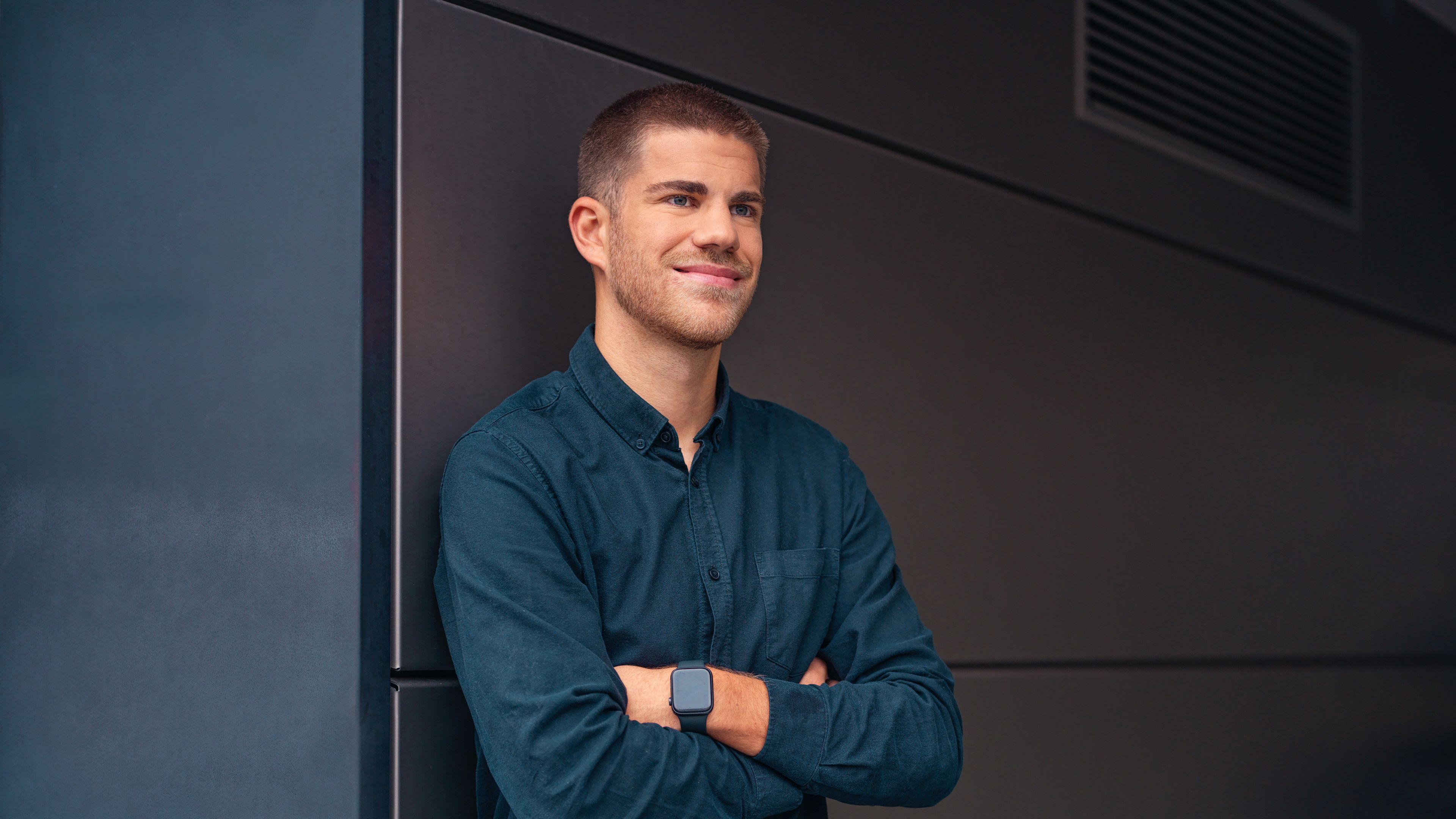 A young man leans against a black wall with his hands in a carbaton.