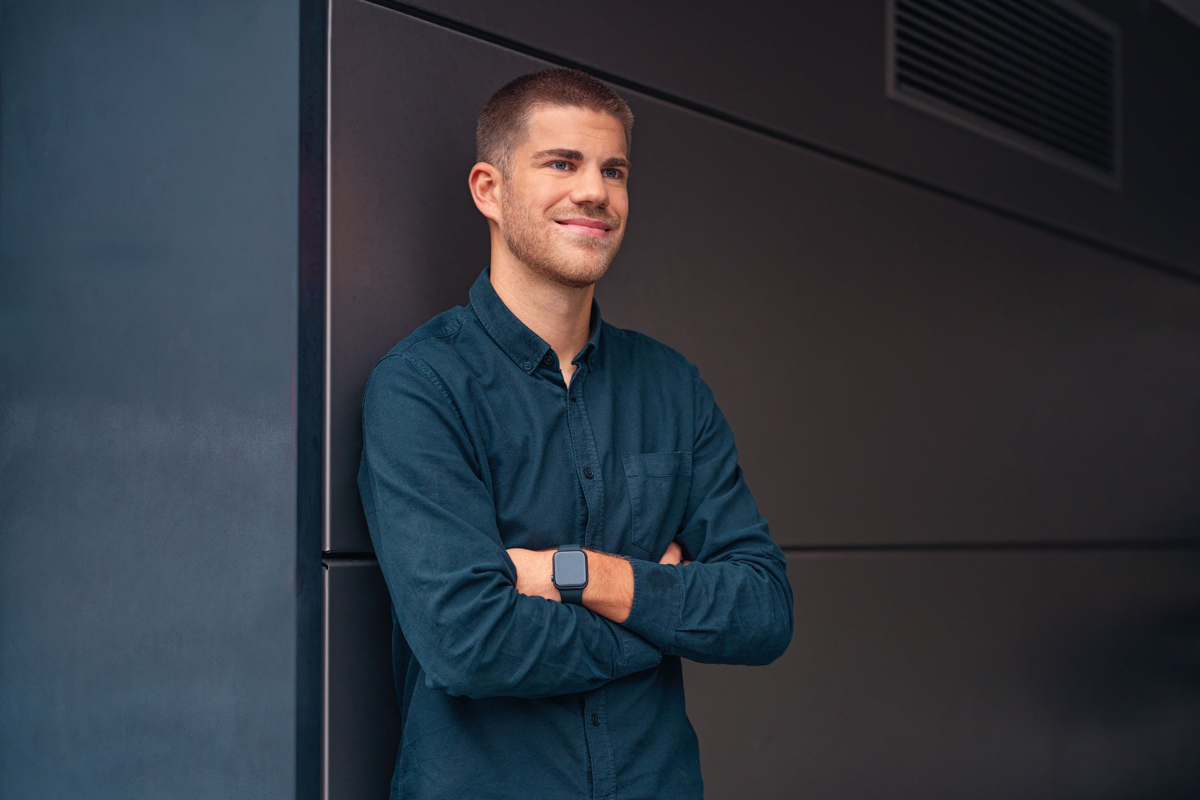 A young man leans against a black wall with his hands in a carbaton.