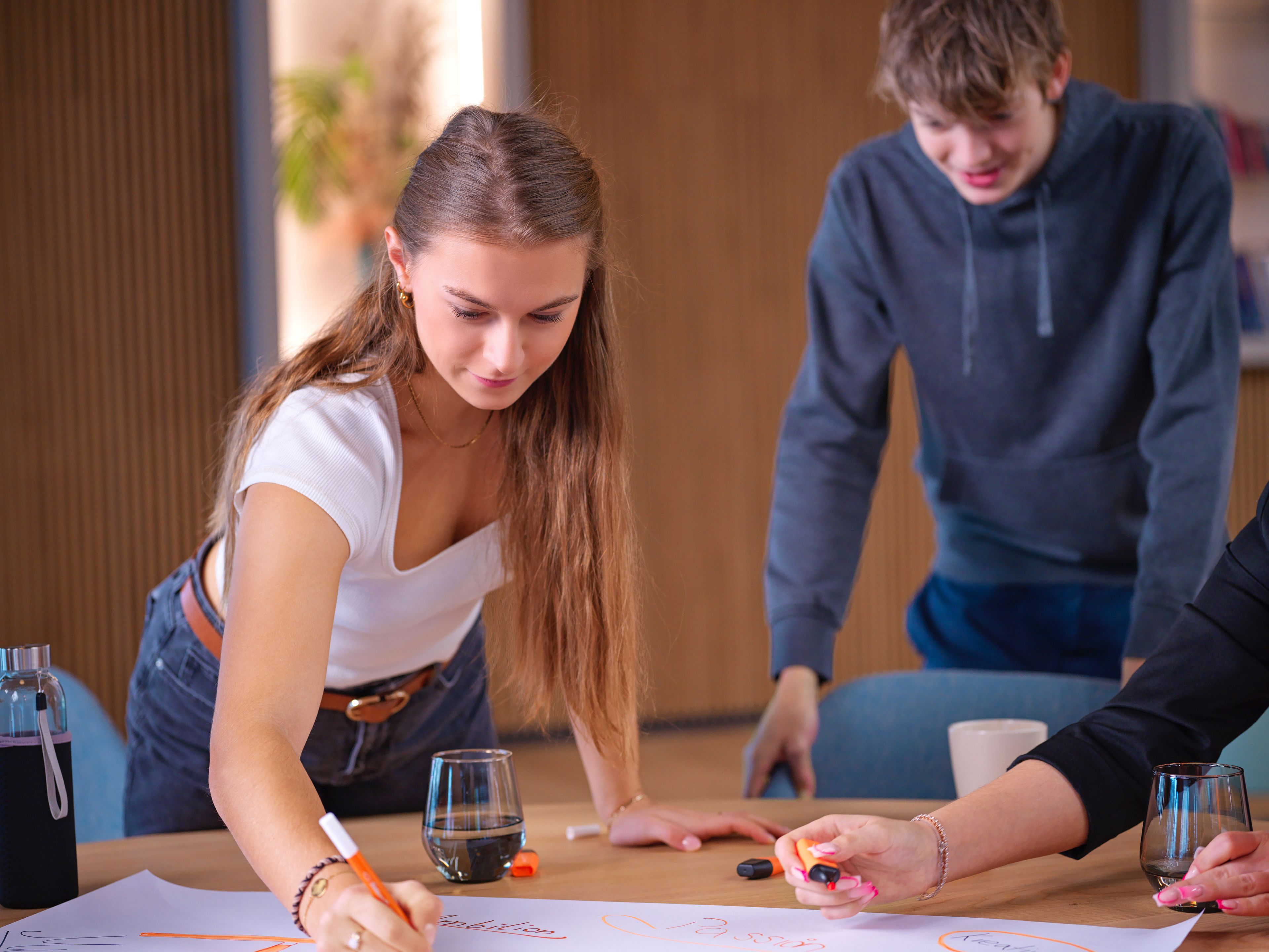 A young girl is writing on a piece of paper with an orange marker, with a young boy behind her.