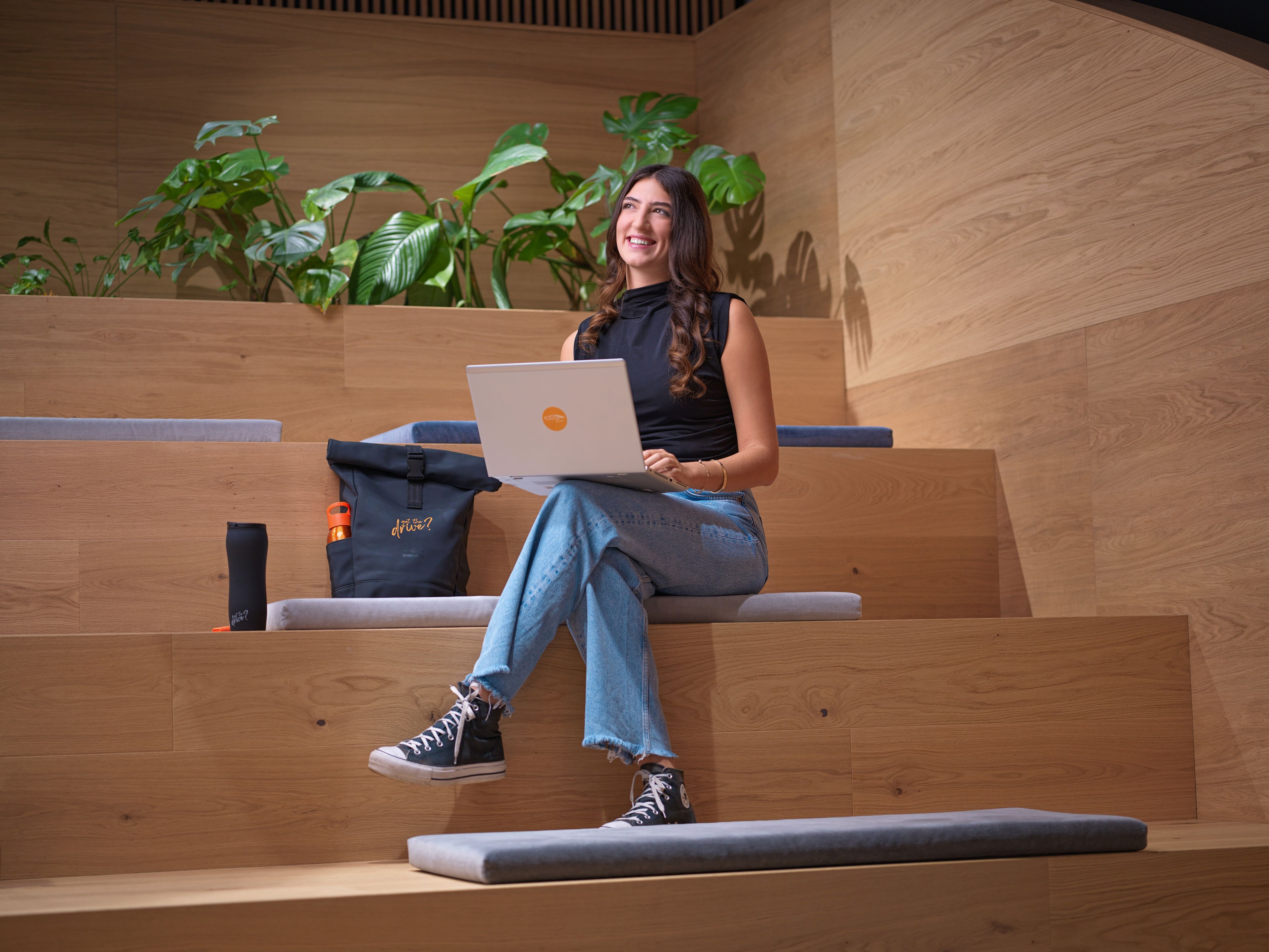 Egy fiatal hölgy ül egy laptoppal egy modern, lépcsőzetes felépítésű előadó teremben, háttérben zöld növényekkel.