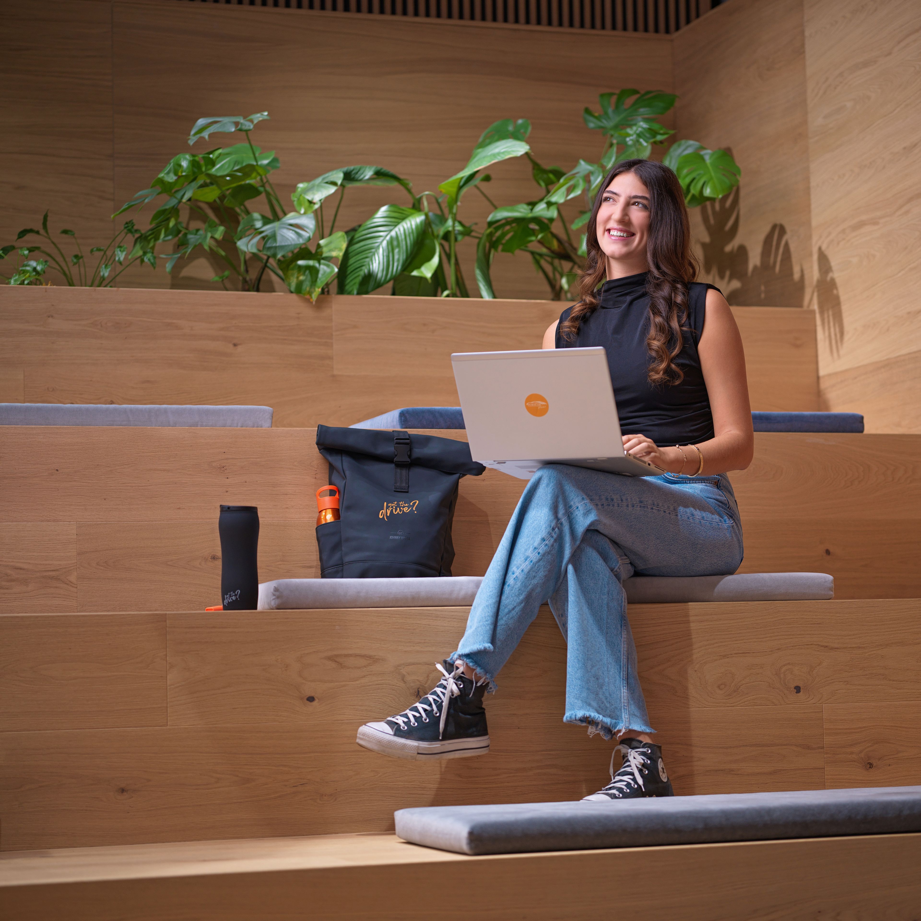 Egy fiatal hölgy ül egy laptoppal egy modern, lépcsőzetes felépítésű előadó teremben, háttérben zöld növényekkel.