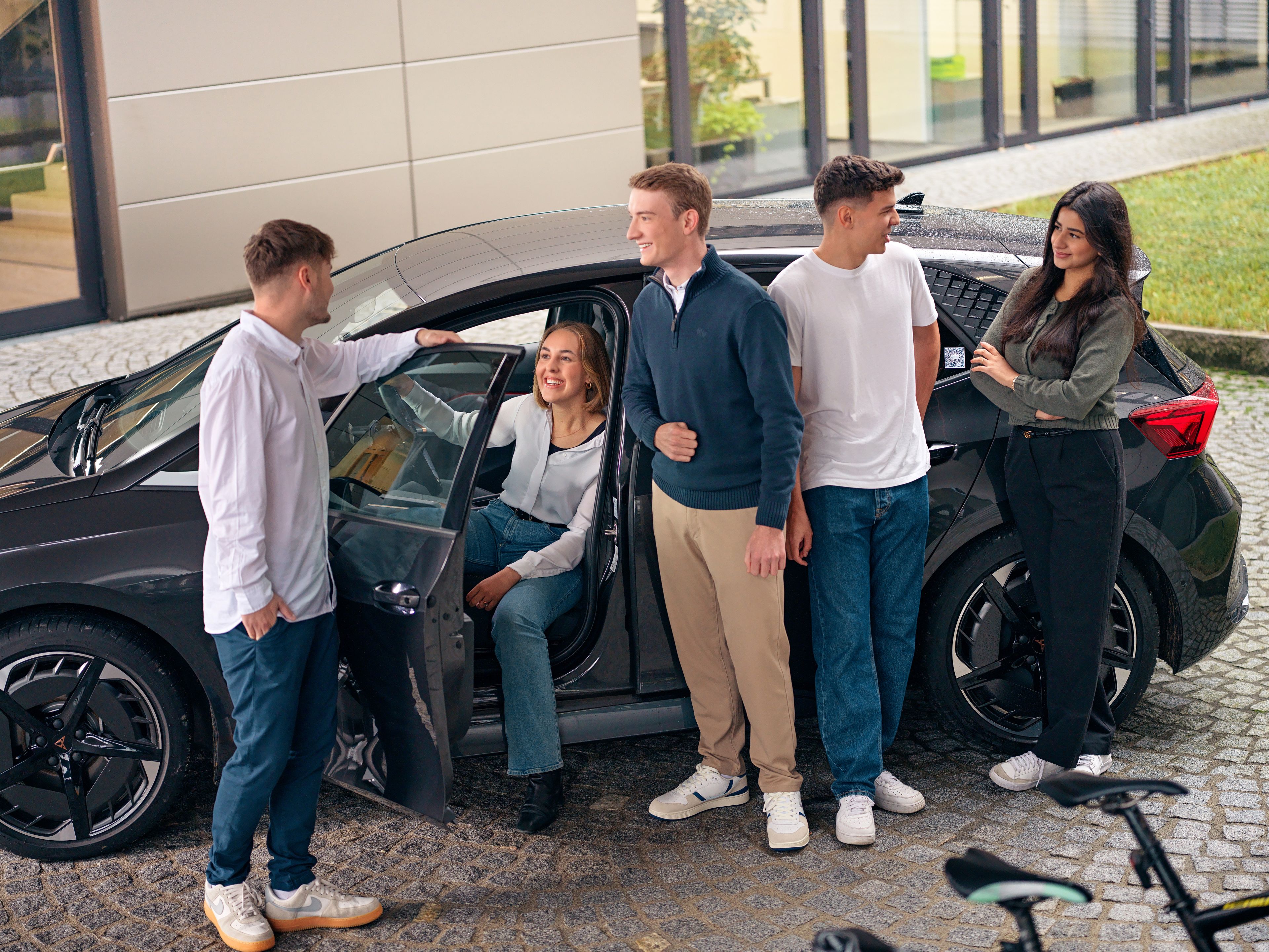 A young lady is sitting in a car, four people are standing next to her, talking.