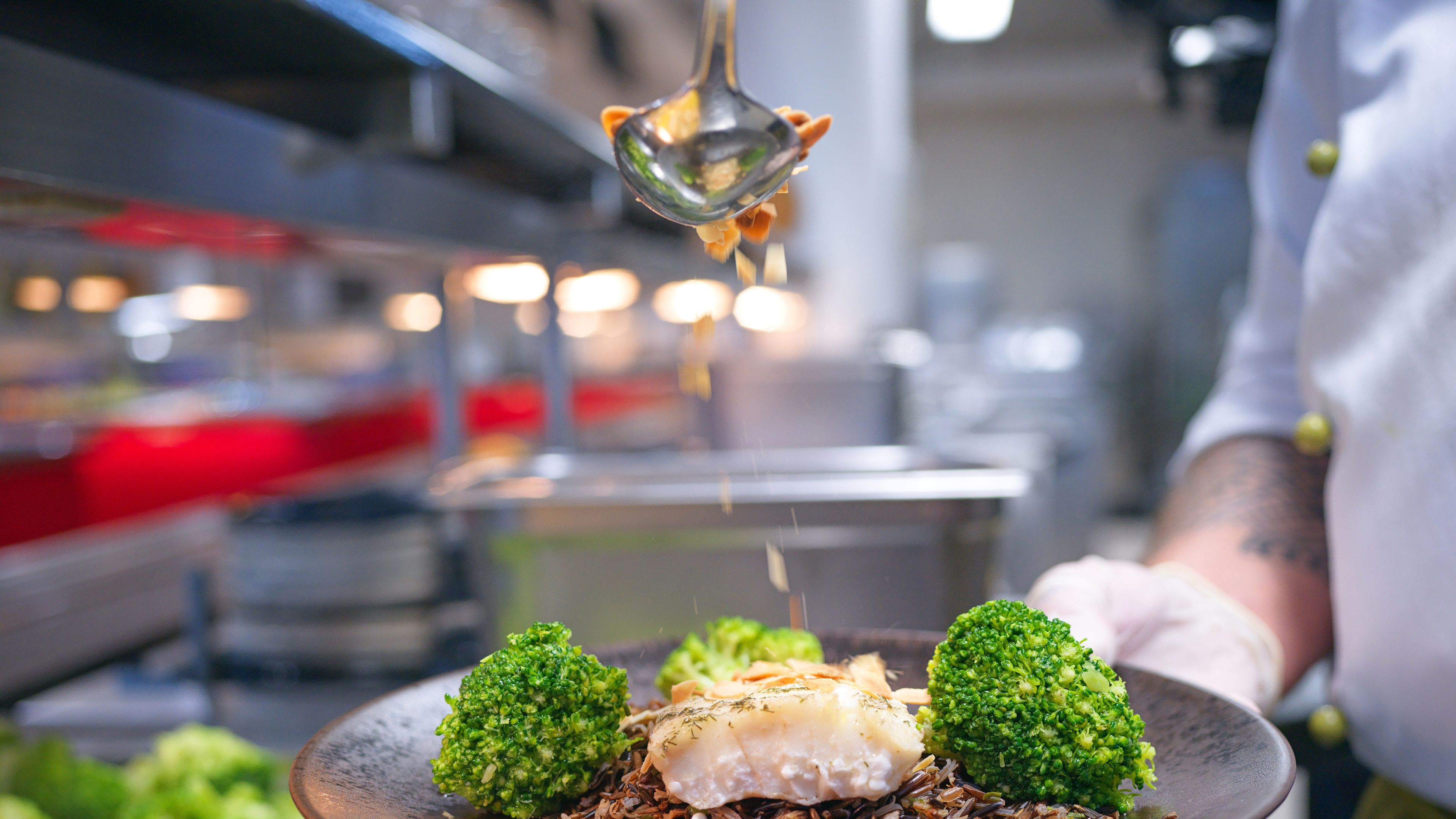 On a plate broccoli, fish and wild rice, on which a chef is sprinkling something.
