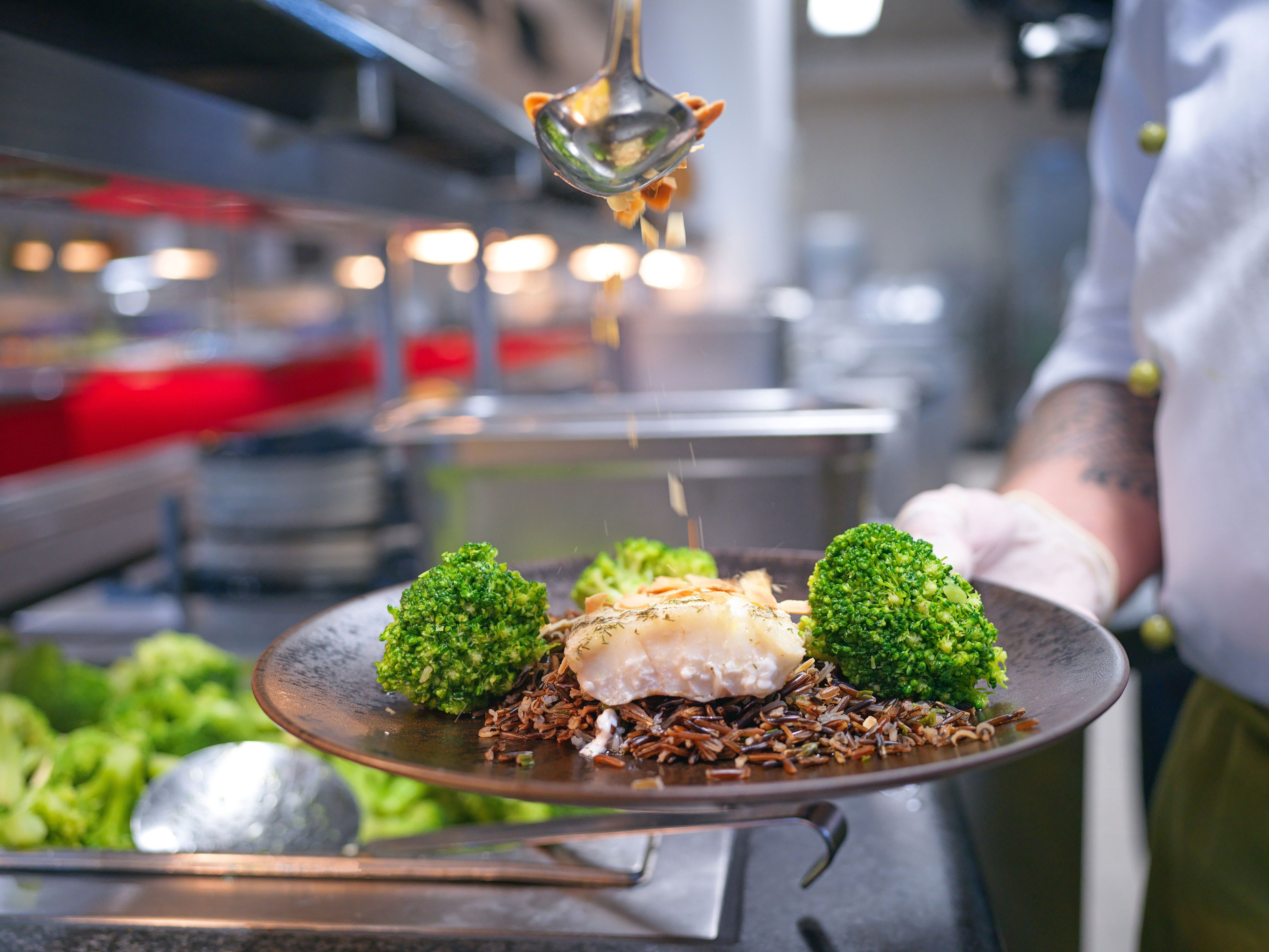 On a plate broccoli, fish and wild rice, on which a chef is sprinkling something.