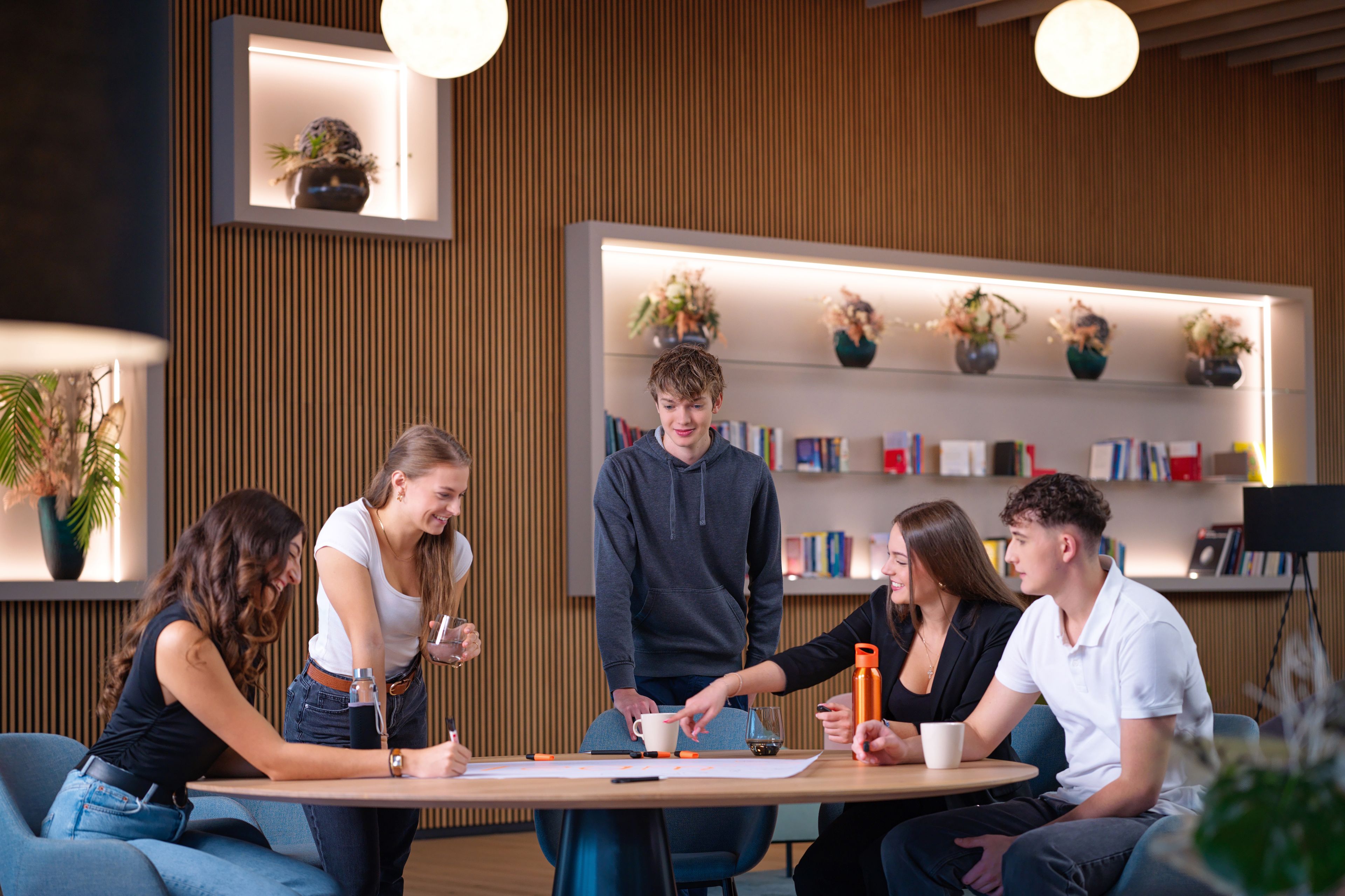 A group of young people sit around a table in a modern office environment and draw.