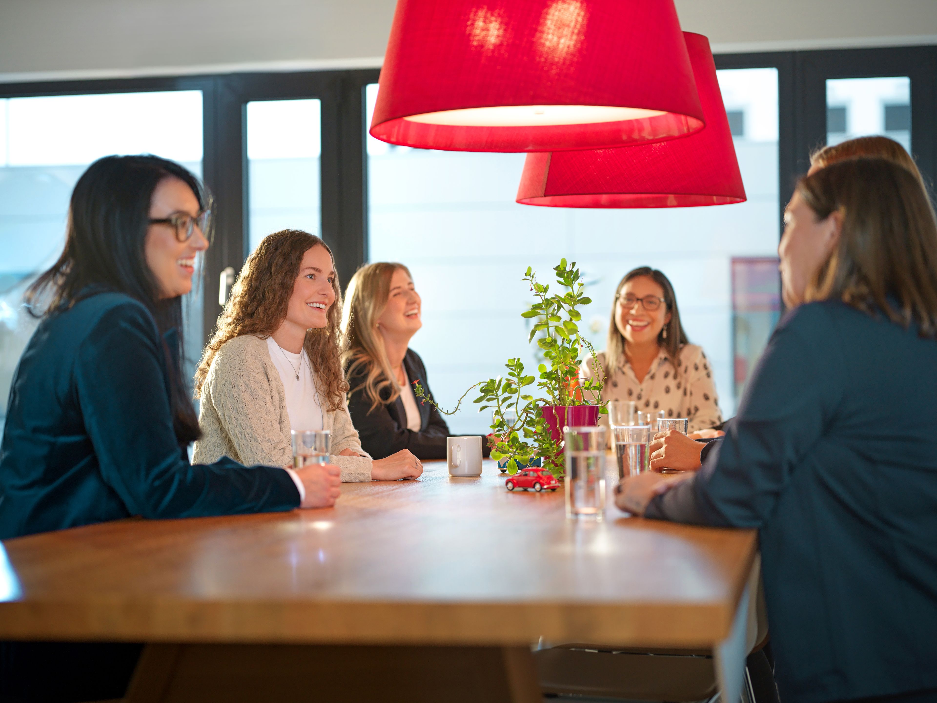 Ladies sit around a table with red lampshades above them and talk.