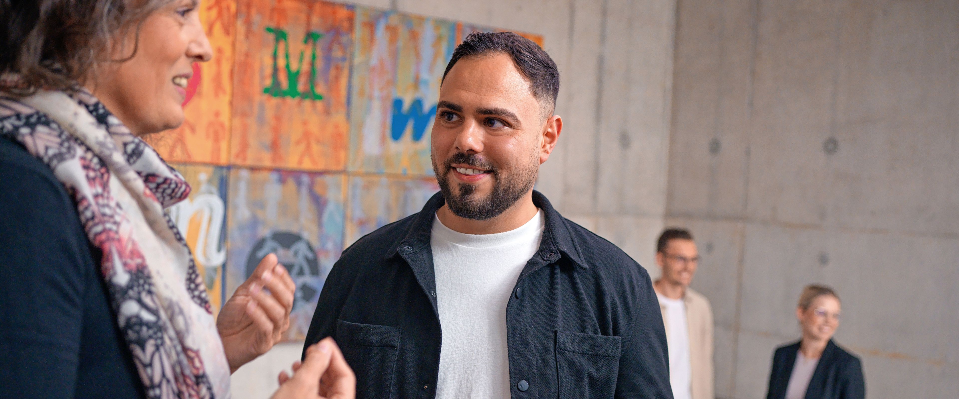 The two of them are talking in a stairwell, behind them is a colorful modern painting.
