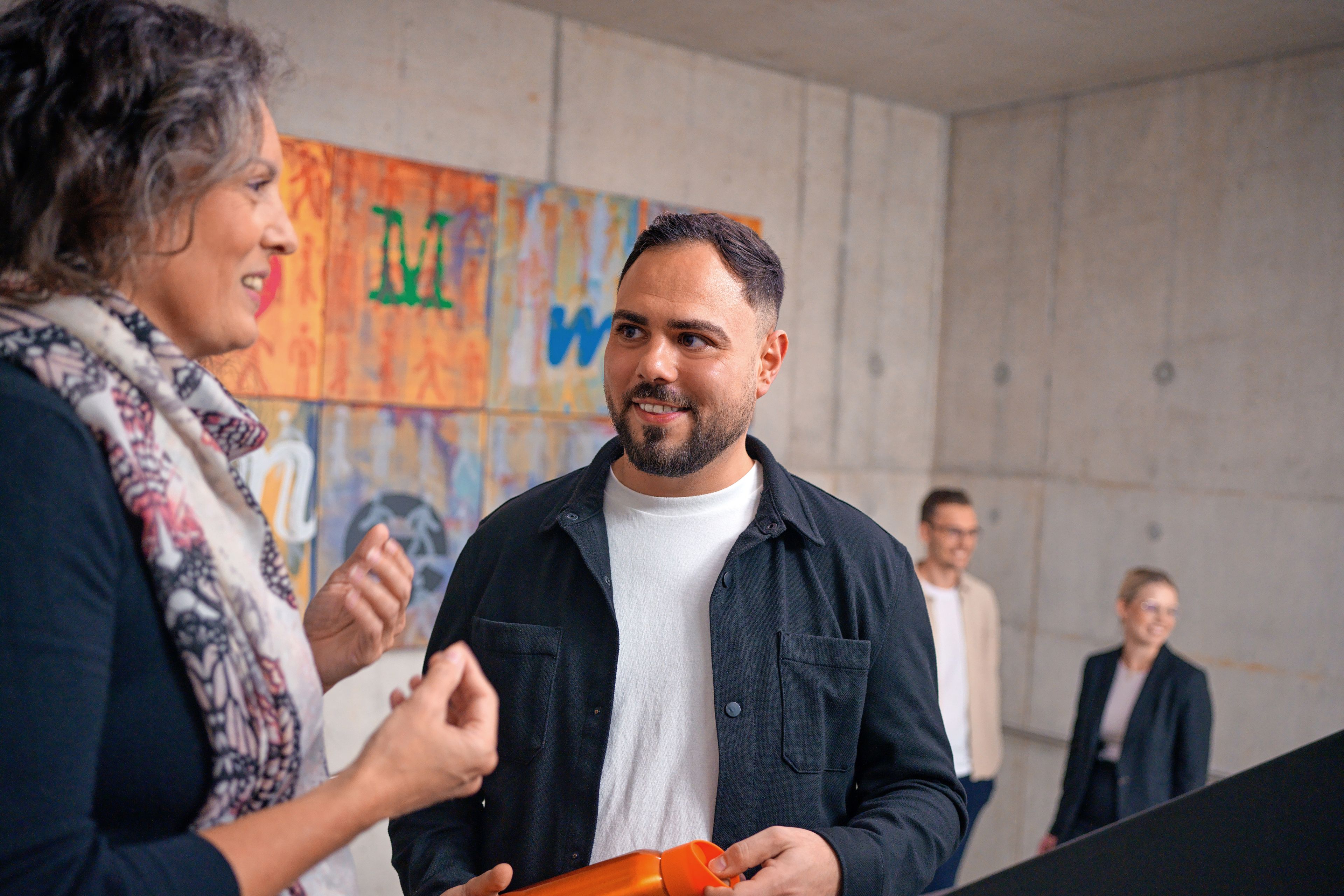 The two of them are talking in a stairwell, behind them is a colorful modern painting.