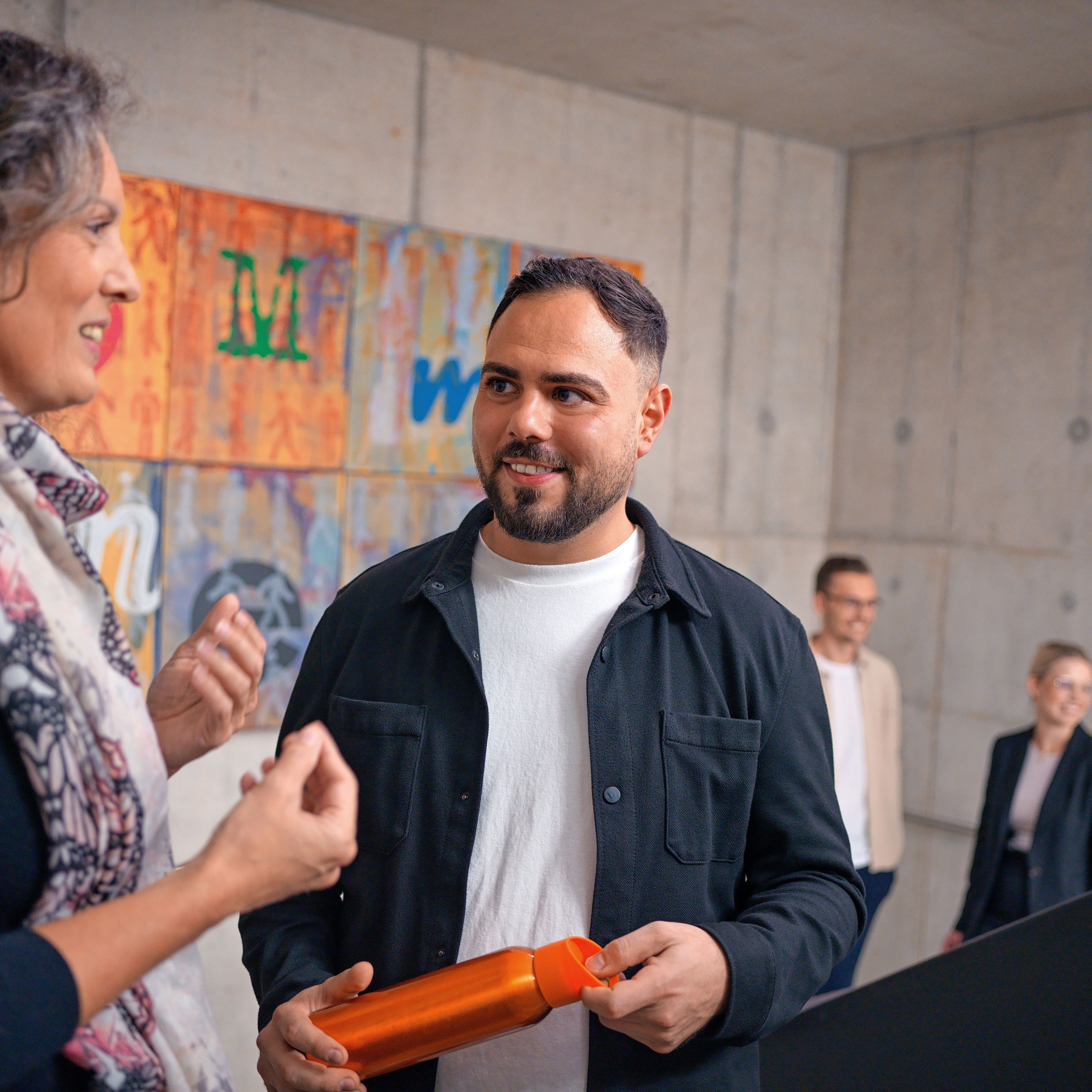 The two of them are talking in a stairwell, behind them is a colorful modern painting.