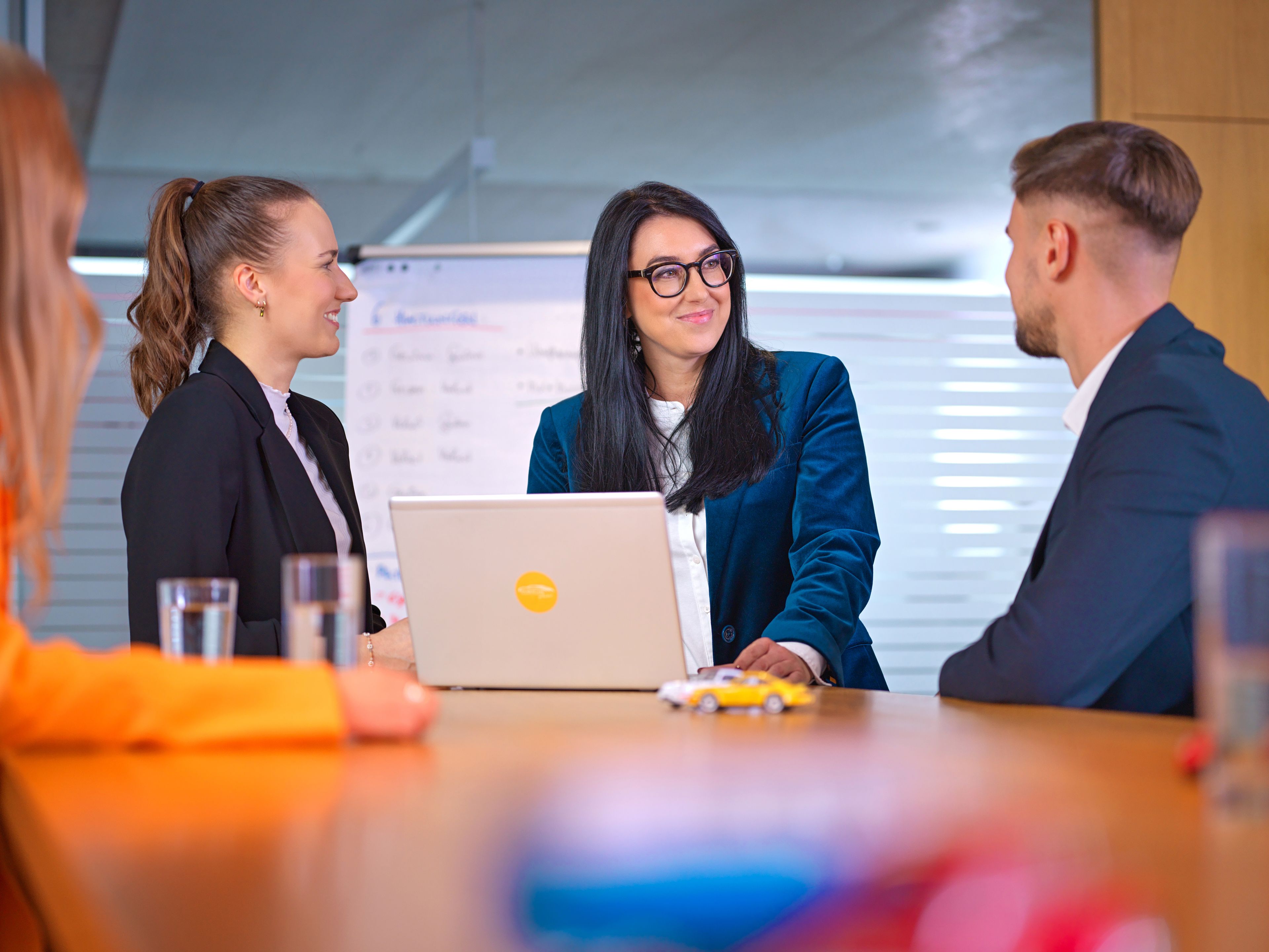 Three people, two ladies and a gentleman, are talking in a café, with black and white photos of old car races behind them.