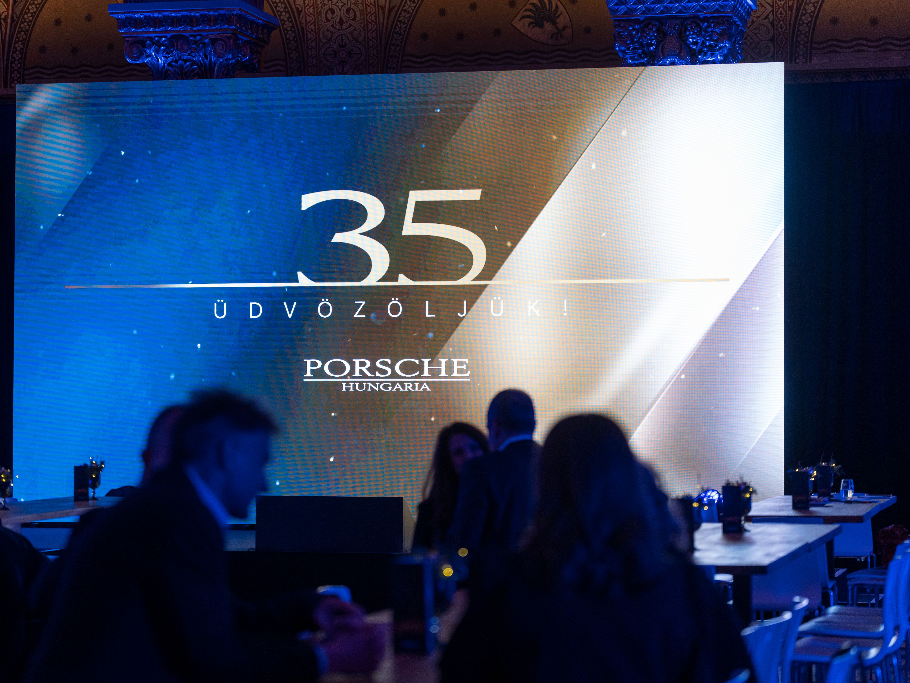 At the 35th birthday celebration of Porsche Hungaria, people sit in front of a festive sign and talk.