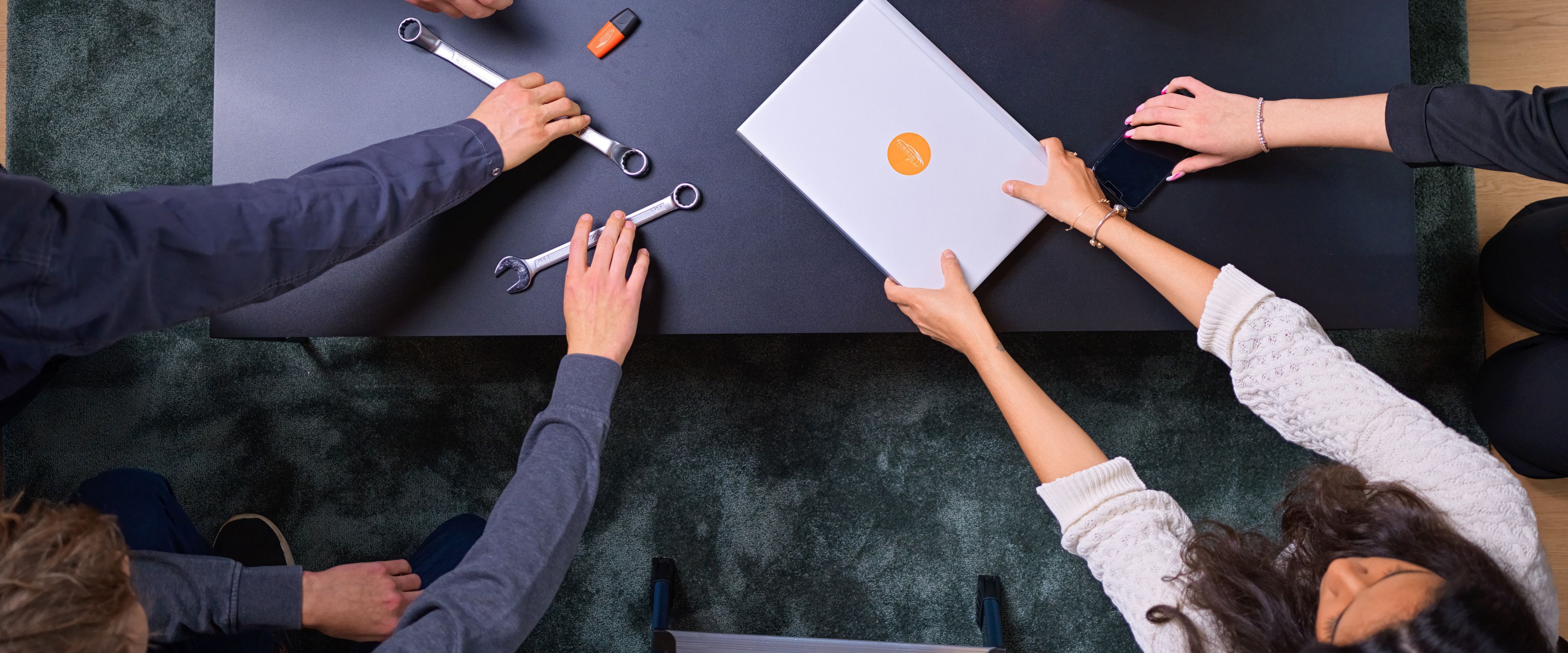 Hands reach over a table and there are tools, a laptop, and other tools in them.
