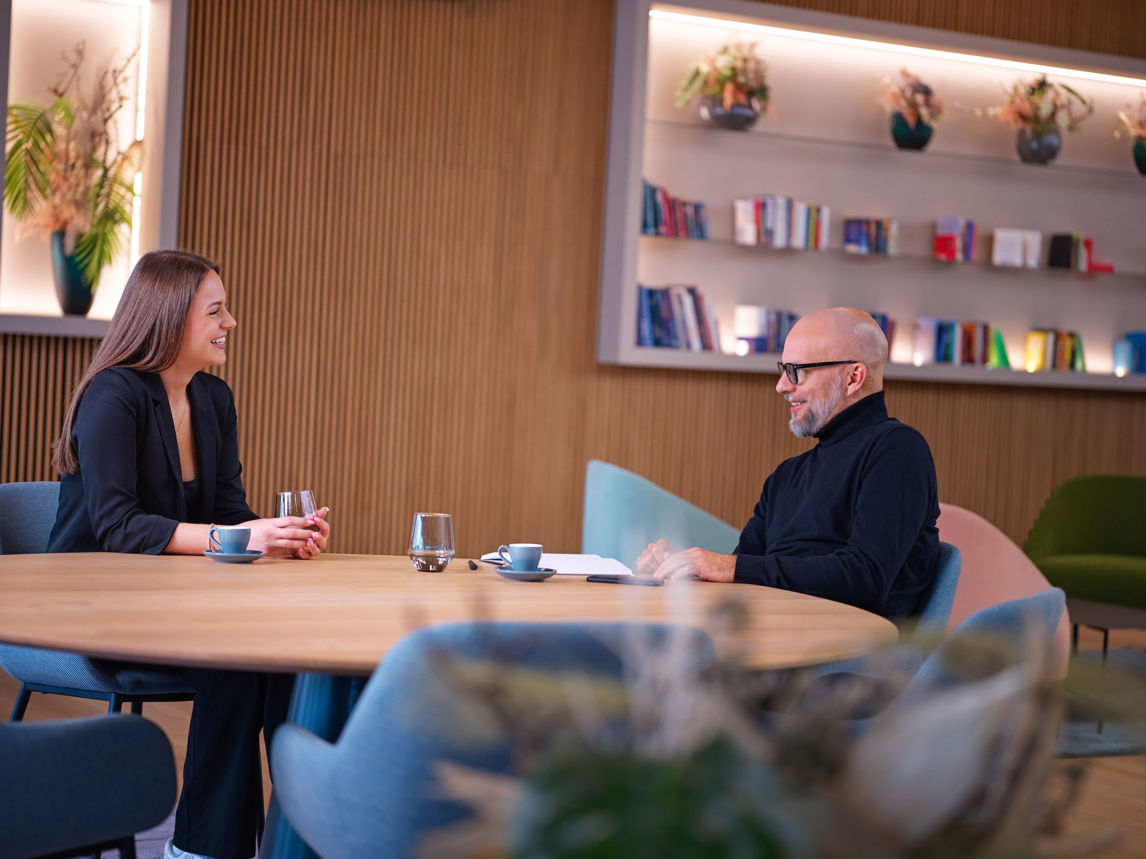 The two of them sit at a table, behind them are books, in front of them is coffee and water.