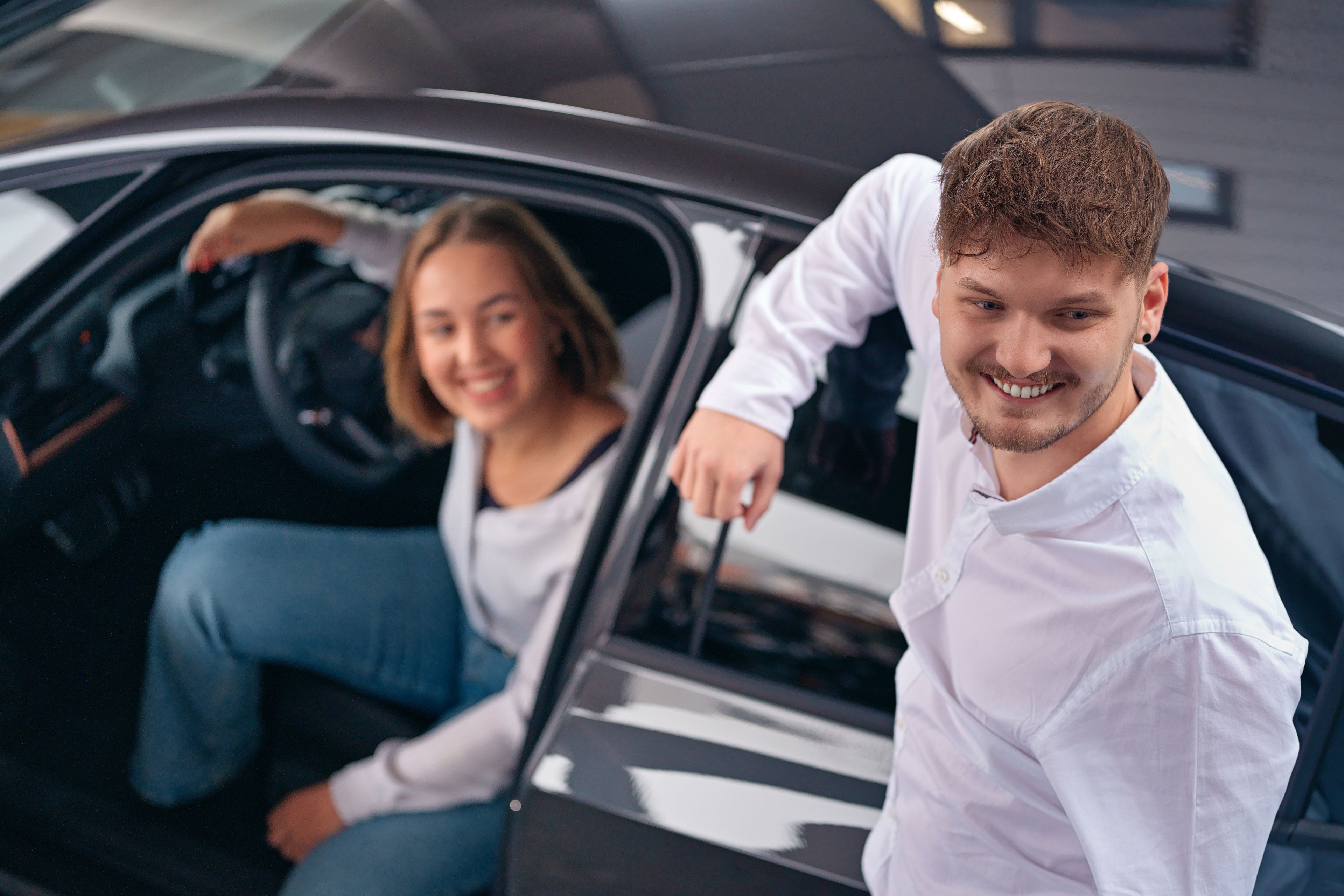 A young girl is sitting in a car with the door open and a young man is elbowing on the car.