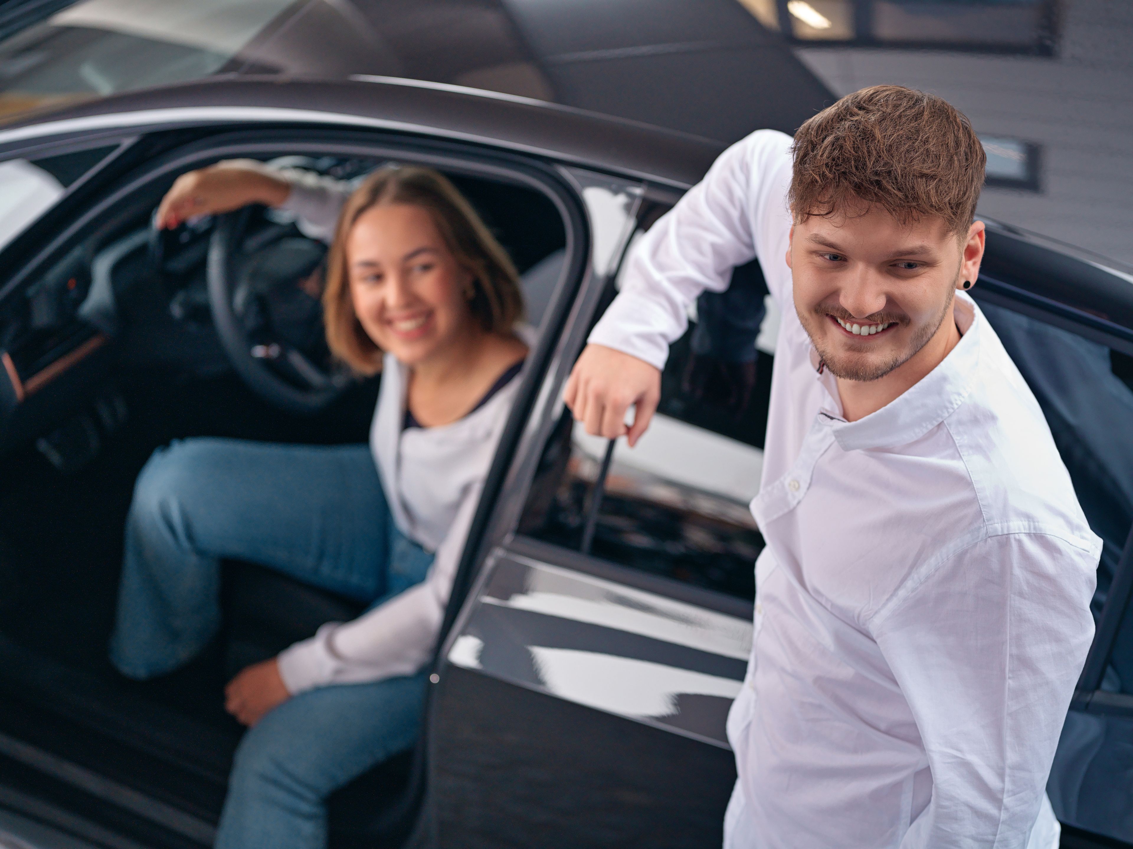 A young girl is sitting in a car with the door open and a young man is elbowing on the car.