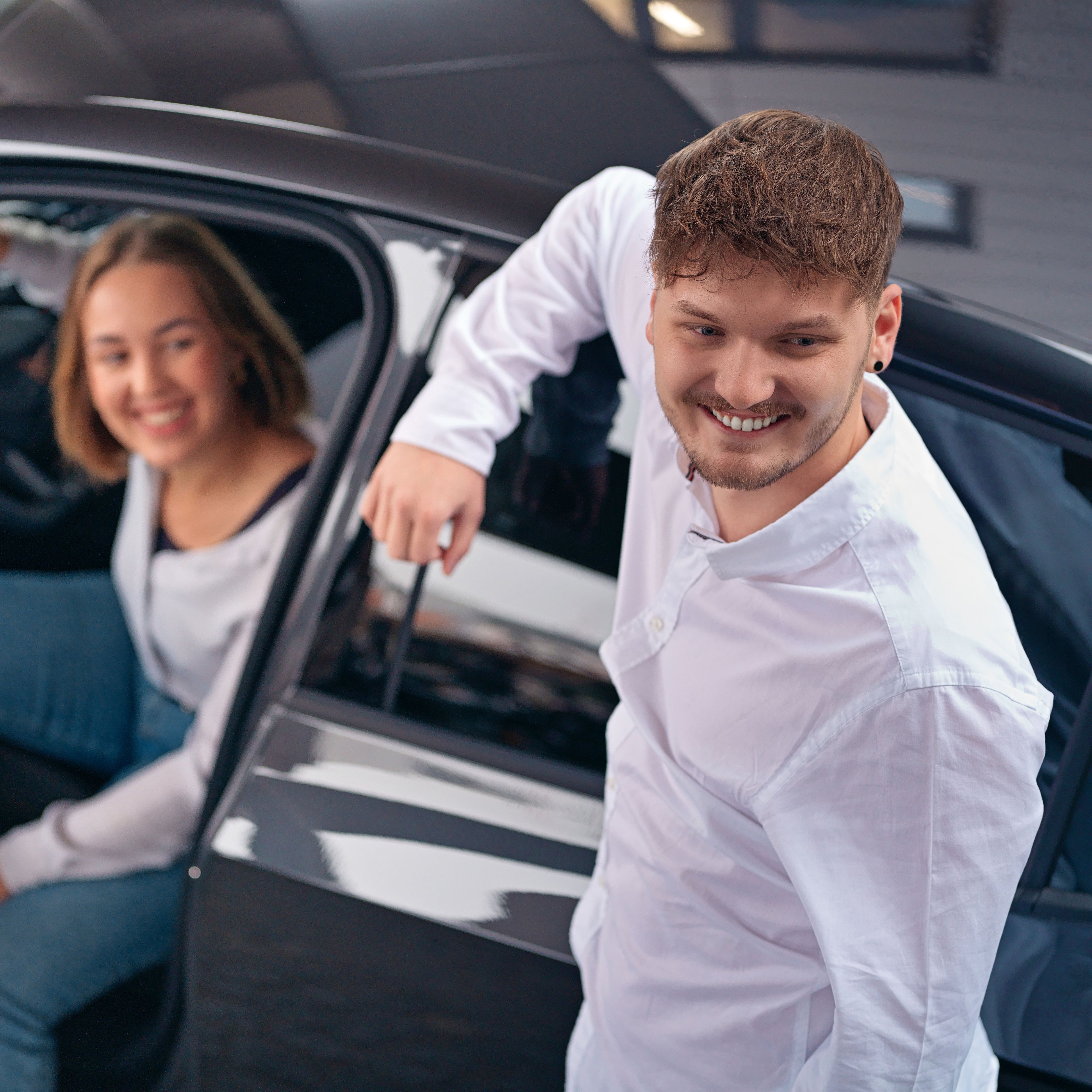 A young girl is sitting in a car with the door open and a young man is elbowing on the car.