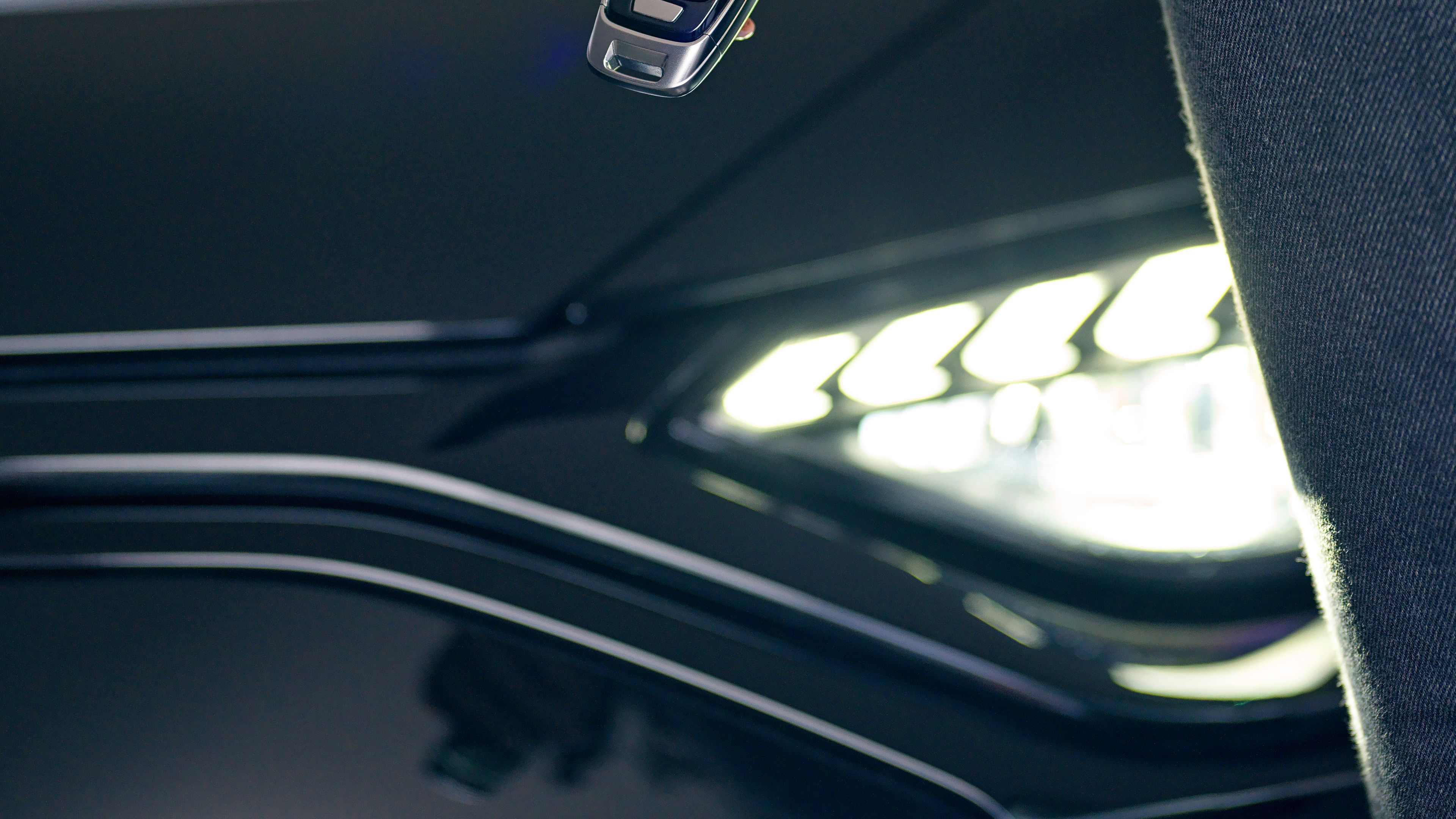 Egy autó nyitó van egy hölgy kezében, mögötte egy fekete autó fényszórója látható.