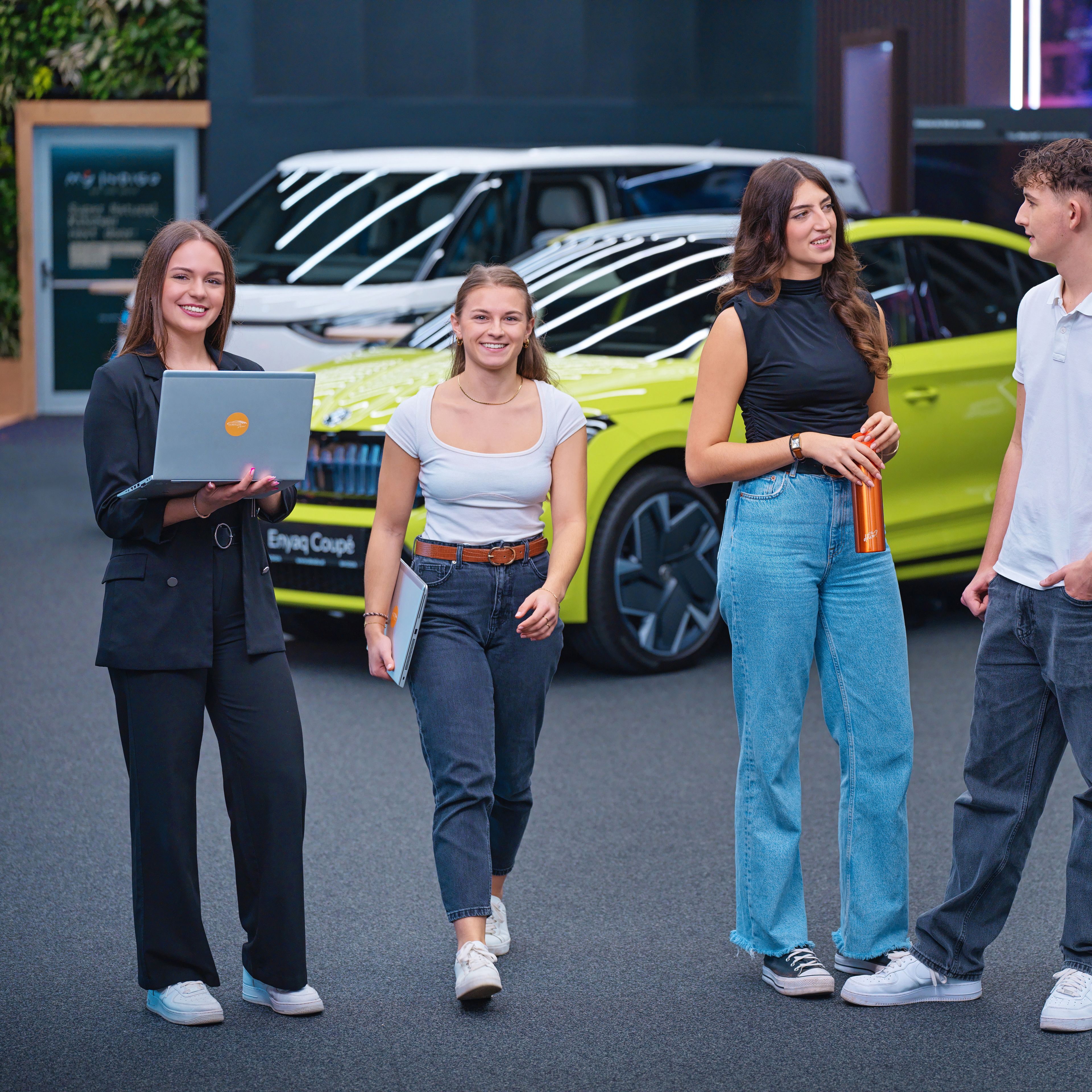 Young people talking, looking at laptops, behind them is a car showroom