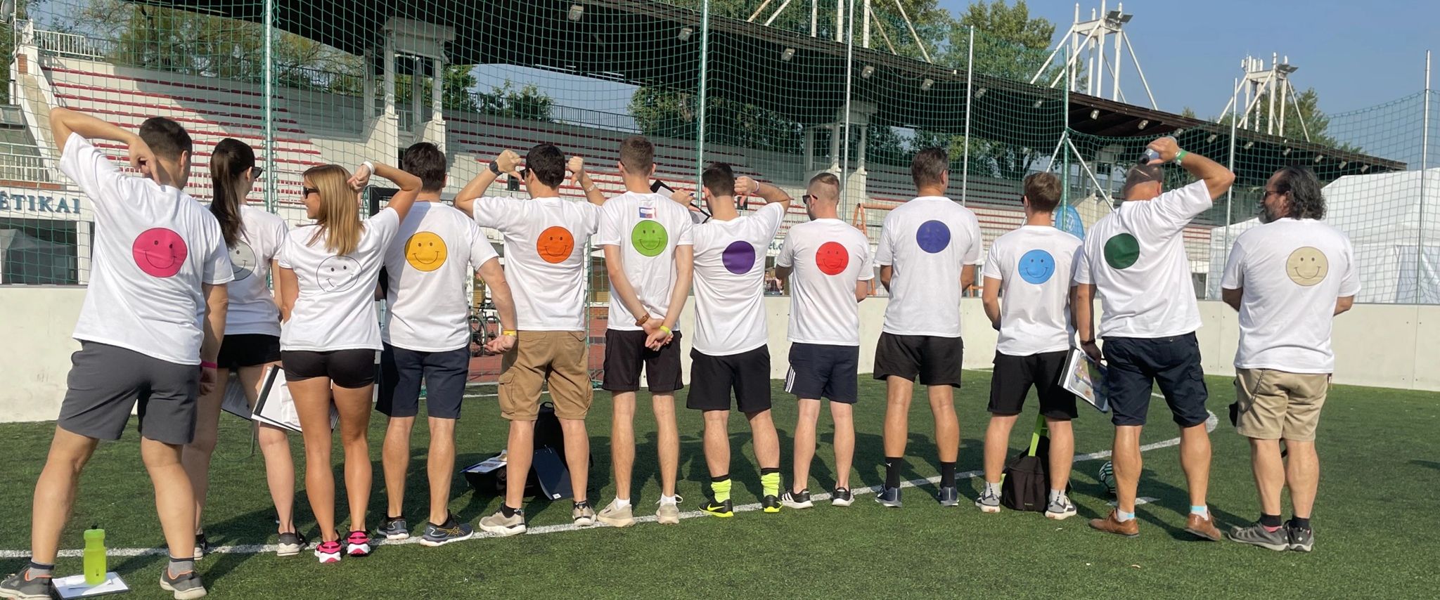 Tizenketten háttal állnak egy sportpályán, pólóikon smiley-k.