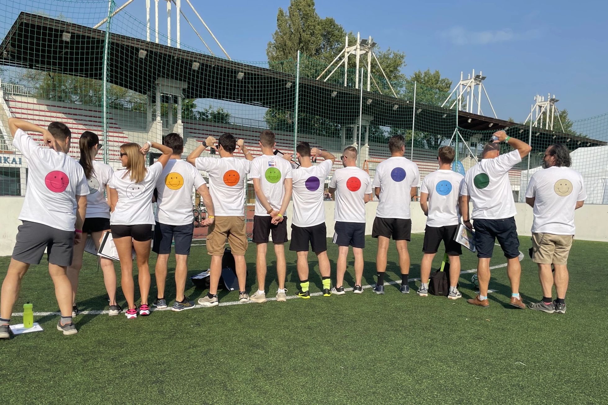 Tizenketten háttal állnak egy sportpályán, pólóikon smiley-k.