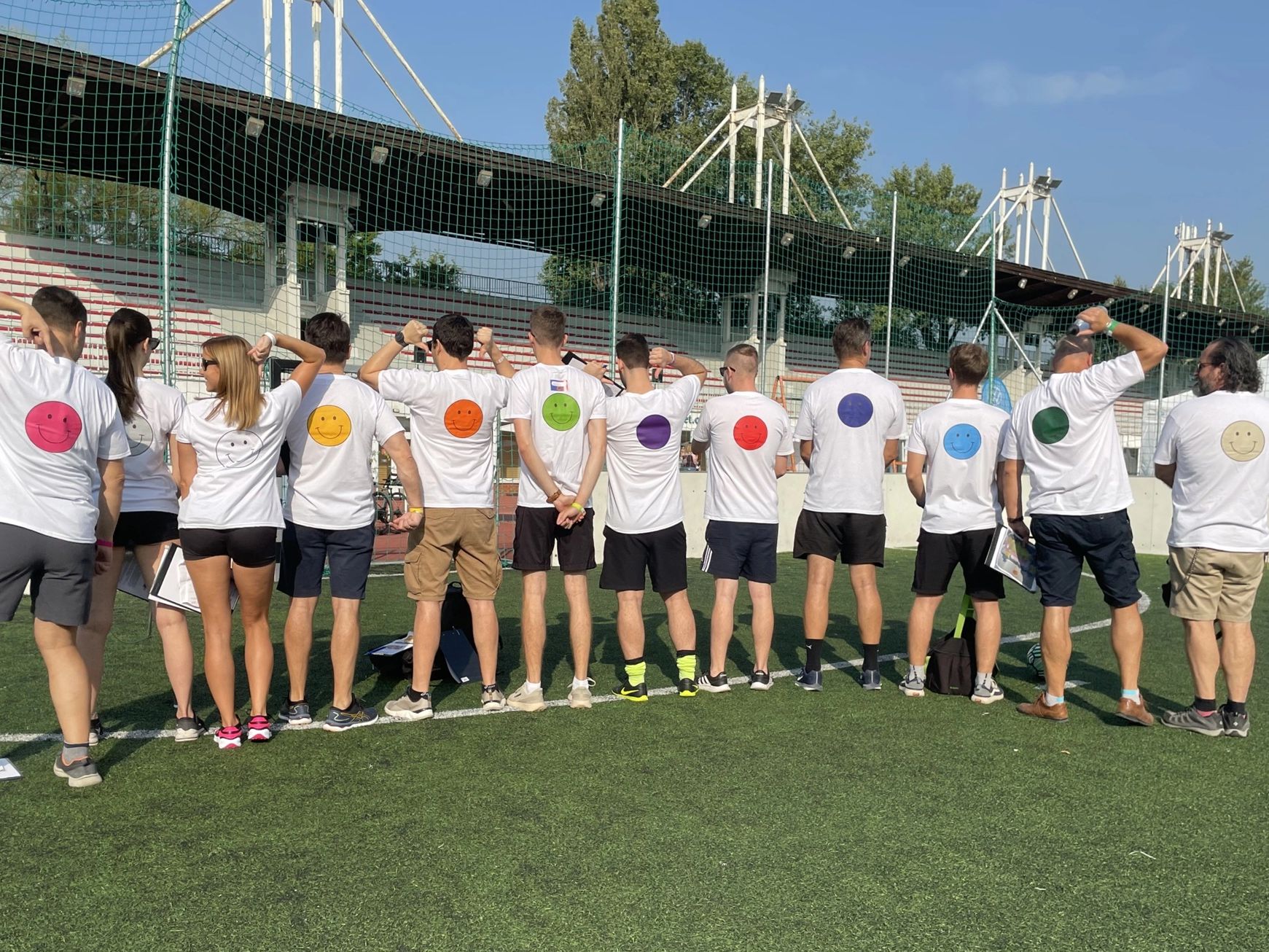 Tizenketten háttal állnak egy sportpályán, pólóikon smiley-k.