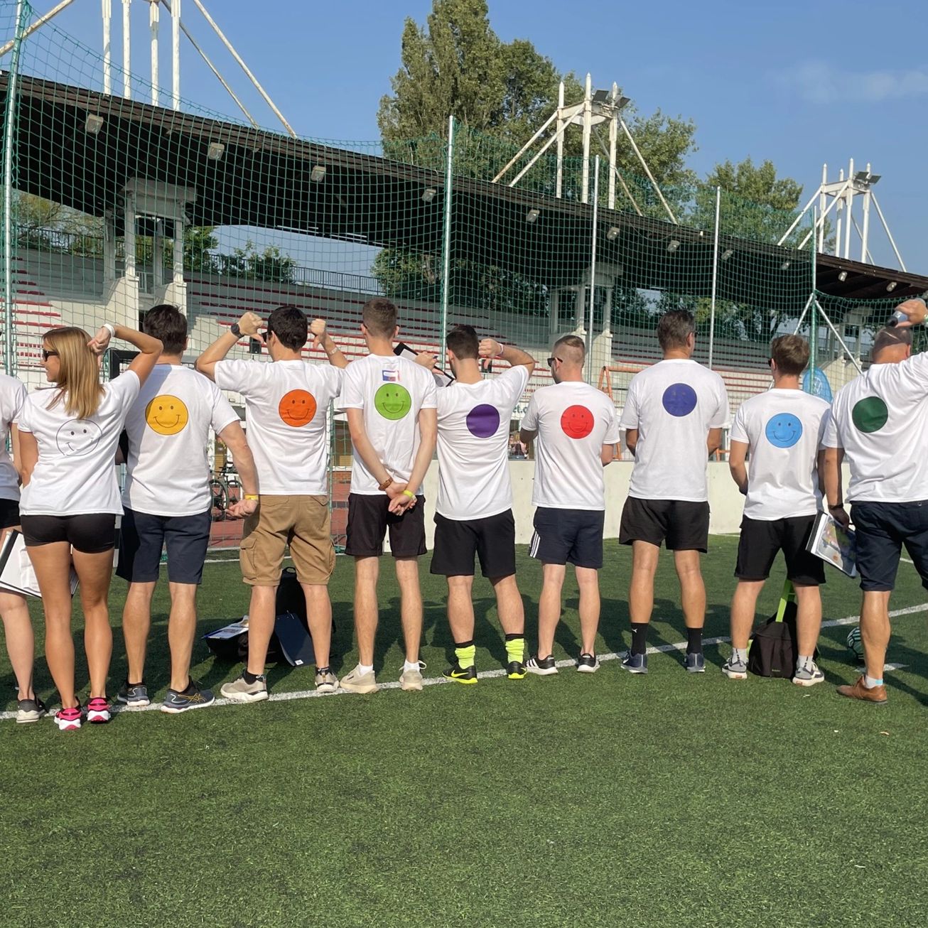 Tizenketten háttal állnak egy sportpályán, pólóikon smiley-k.